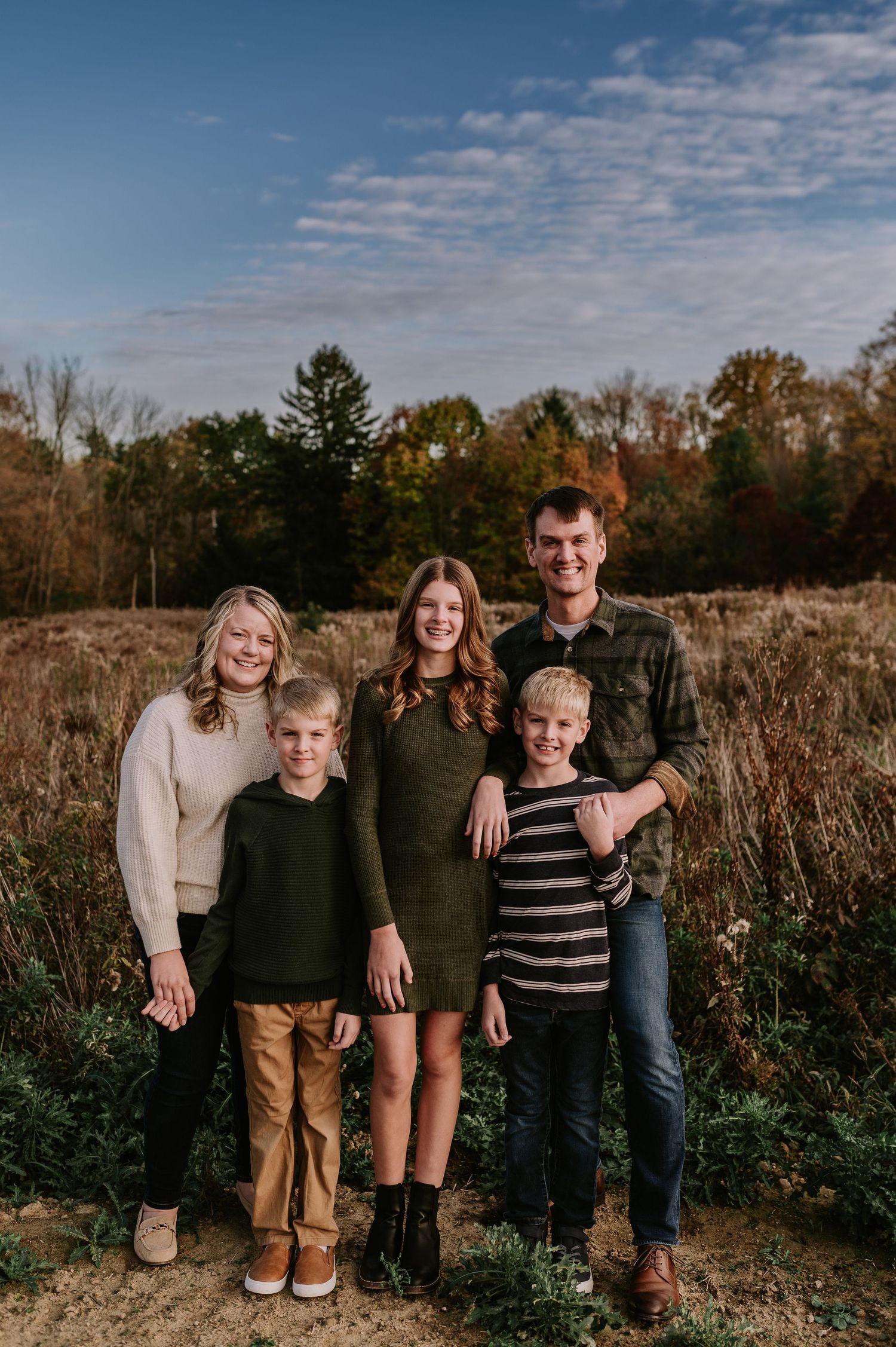 Family of five poses together in a rustic field with autumn trees in the background.