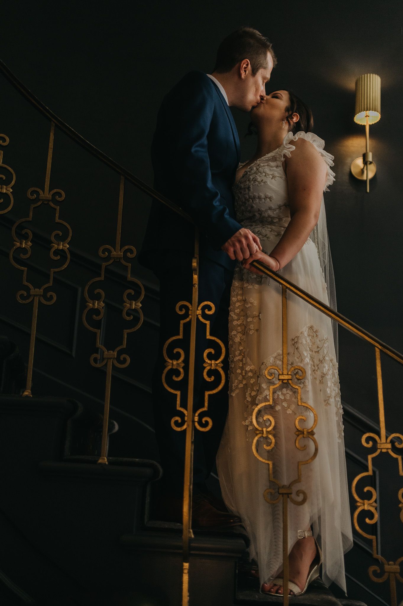 Bride and groom share an intimate moment on ornate golden staircase with vintage lighting.