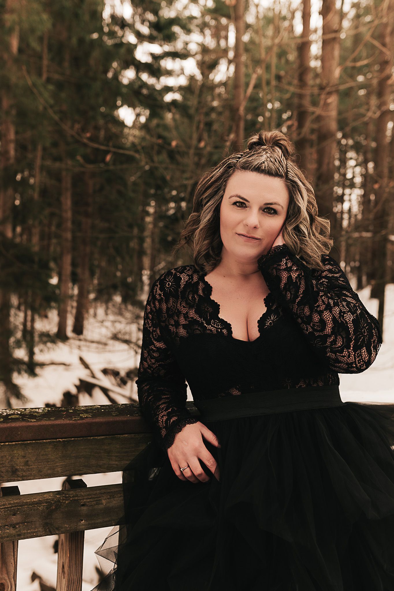 A woman in black lace gown poses playfully in snowy winter forest setting.