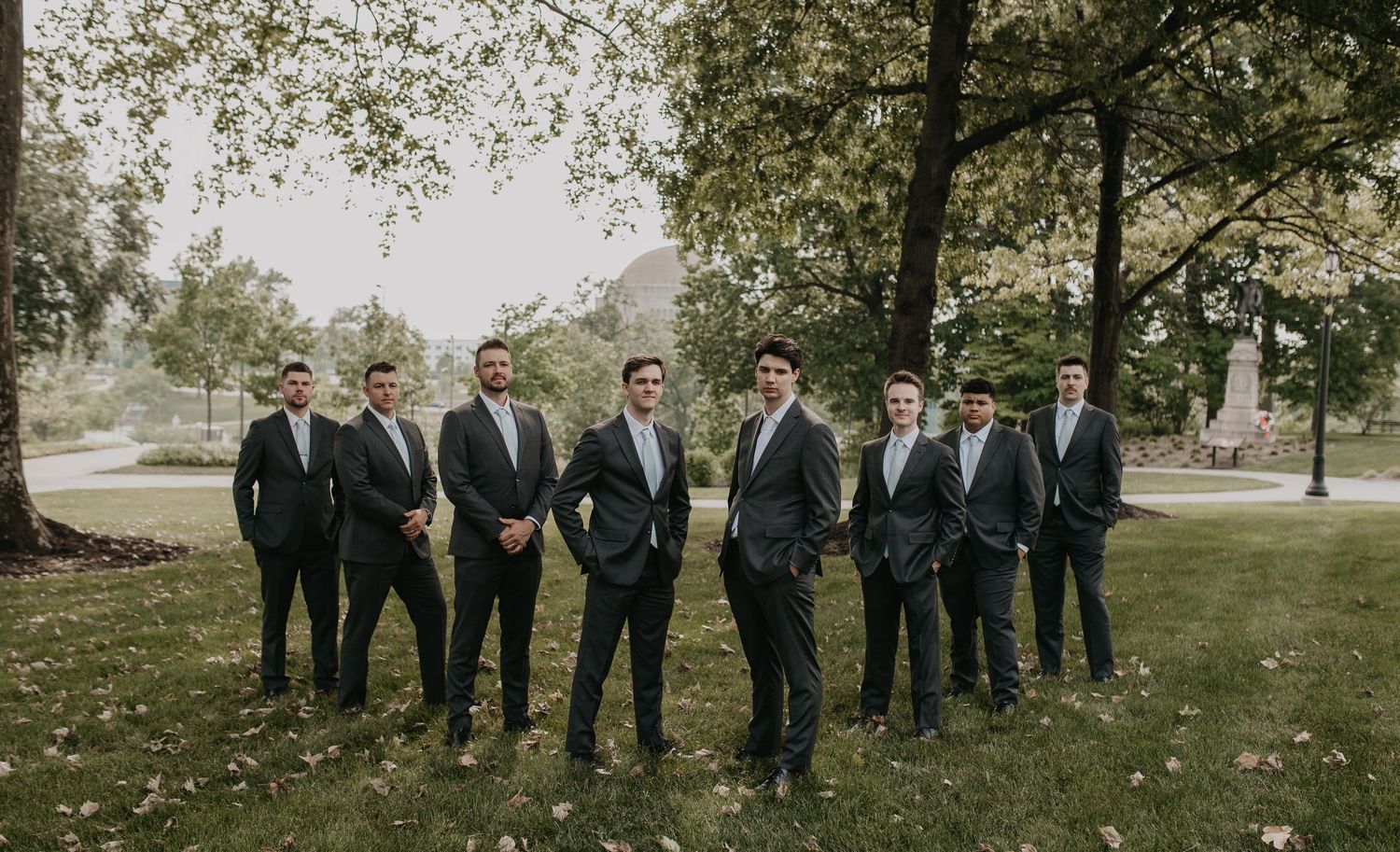 A group of groomsmen in dark suits pose together on a lawn beneath trees for a wedding photo.