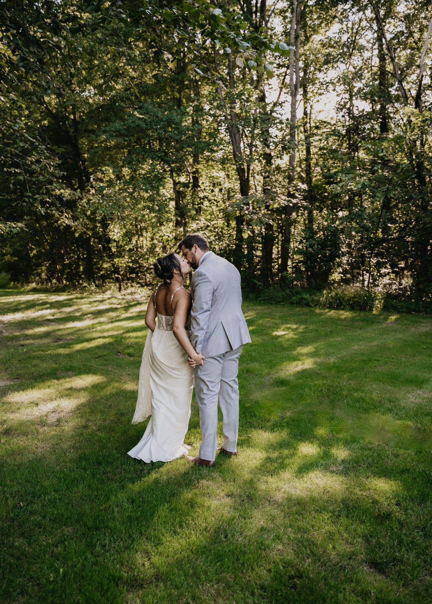 Wedding couple walking together on lush green grass with sunlit trees in the background.