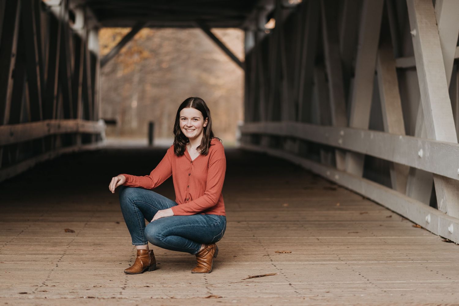 A casual portrait of someone in an orange sweater and jeans crouching on a covered wooden bridge.
