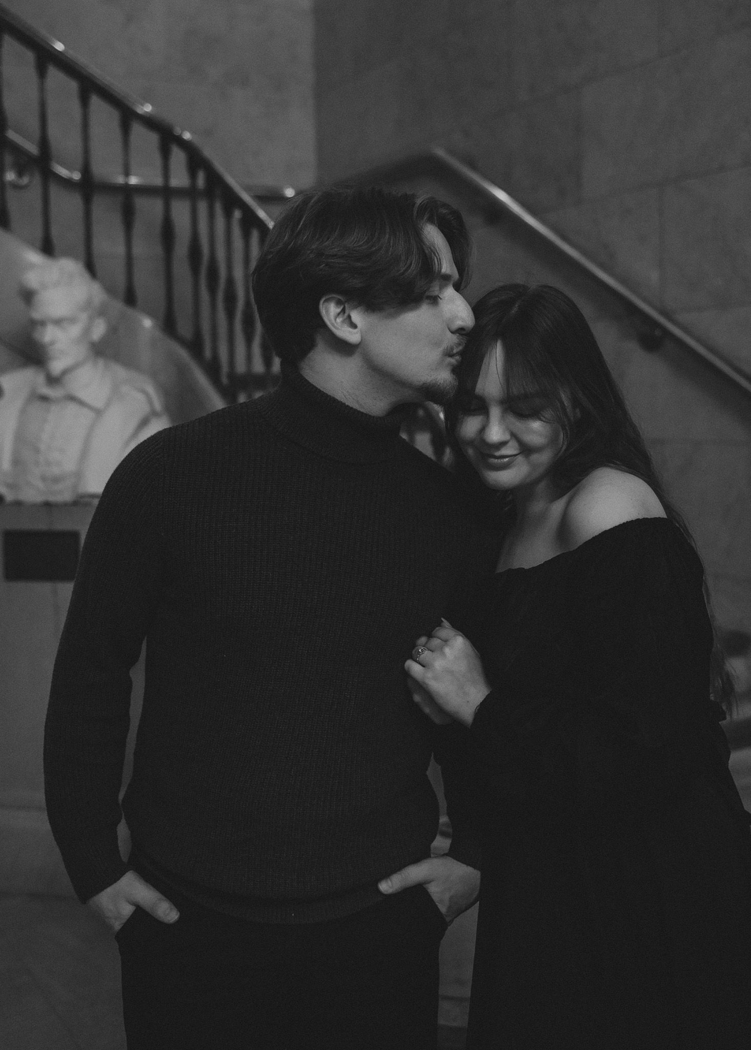 A romantic couple in black clothing share intimate moments in a dimly lit stairwell setting.