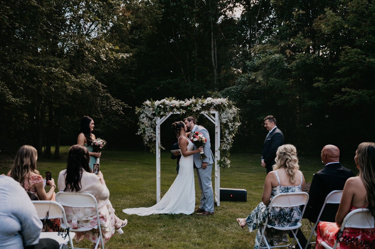 Wedding guests gather outdoors for a casual summer ceremony in a lush green park setting.