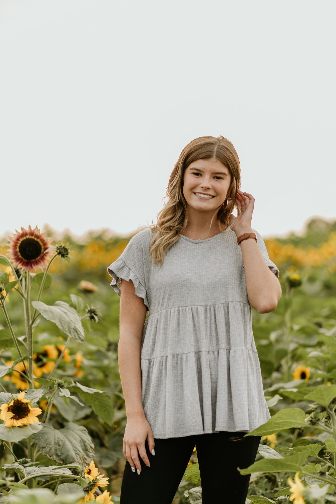 Young woman in a casual gray ruffle top poses cheerfully in a blooming sunflower field on a bright sunny day.