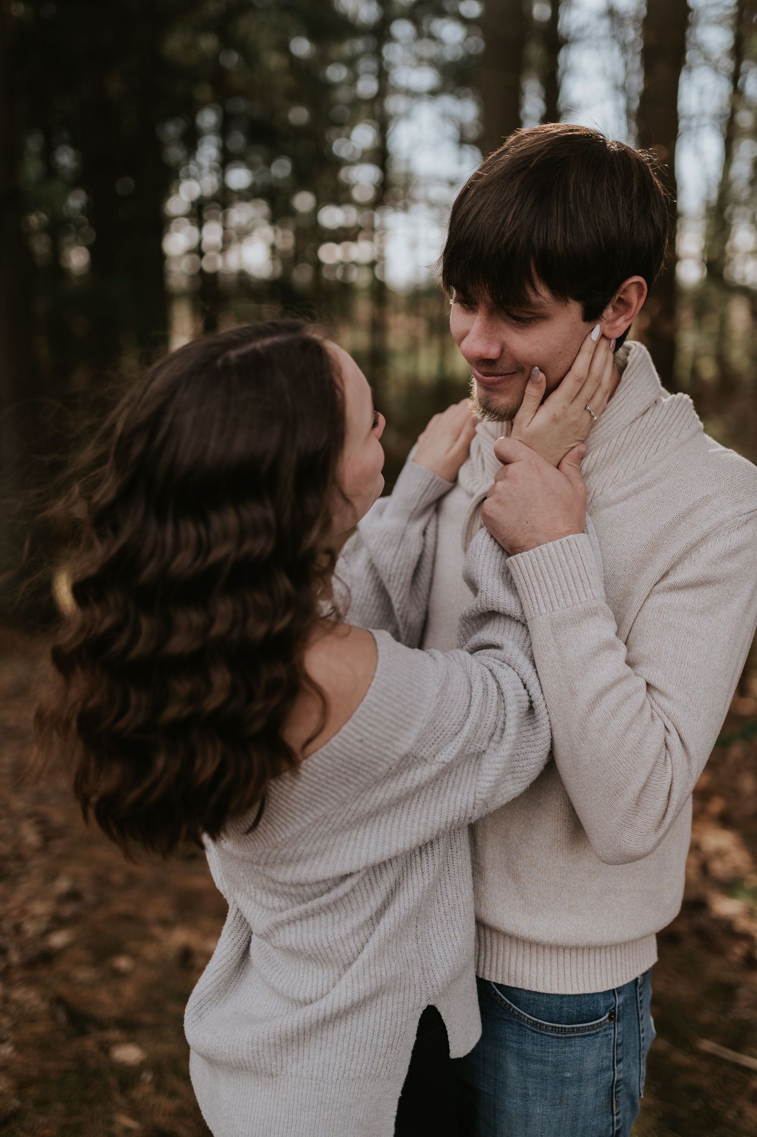 An engaged couple in neutral sweaters share candid laughter and embraces in a wooded setting.