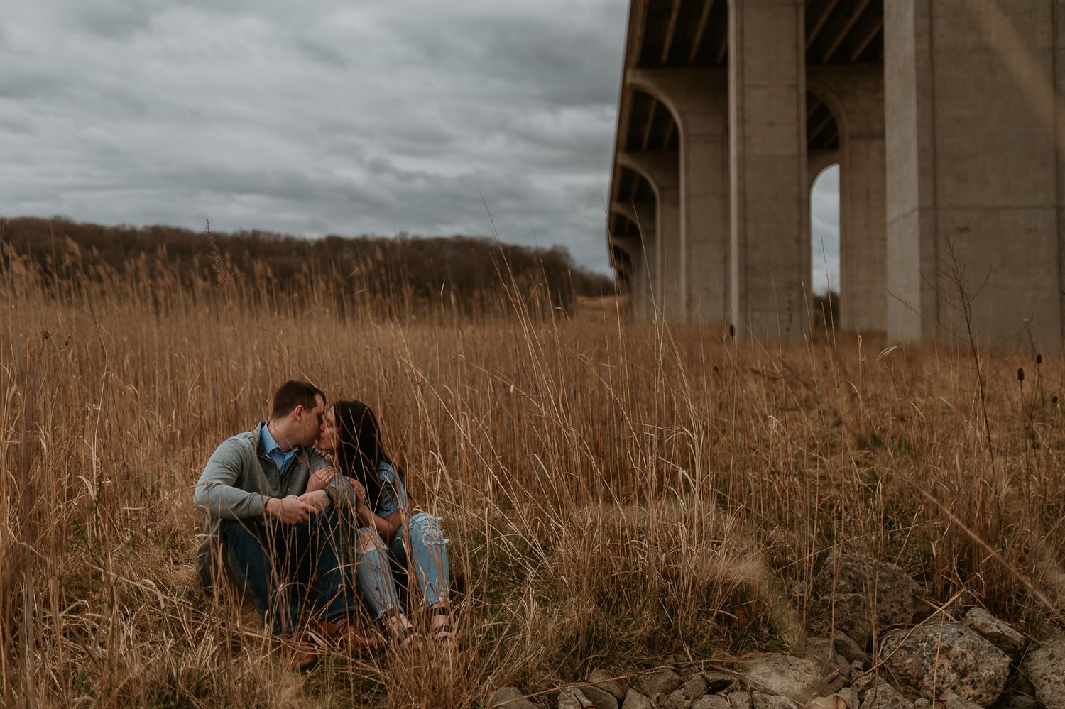 A couple explores a grassy field beneath towering concrete bridge pillars during golden hour.