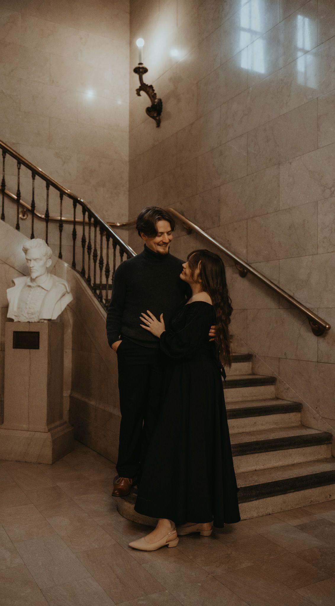 A couple in elegant black attire share an intimate moment on a grand marble staircase with a classical bust sculpture nearby.