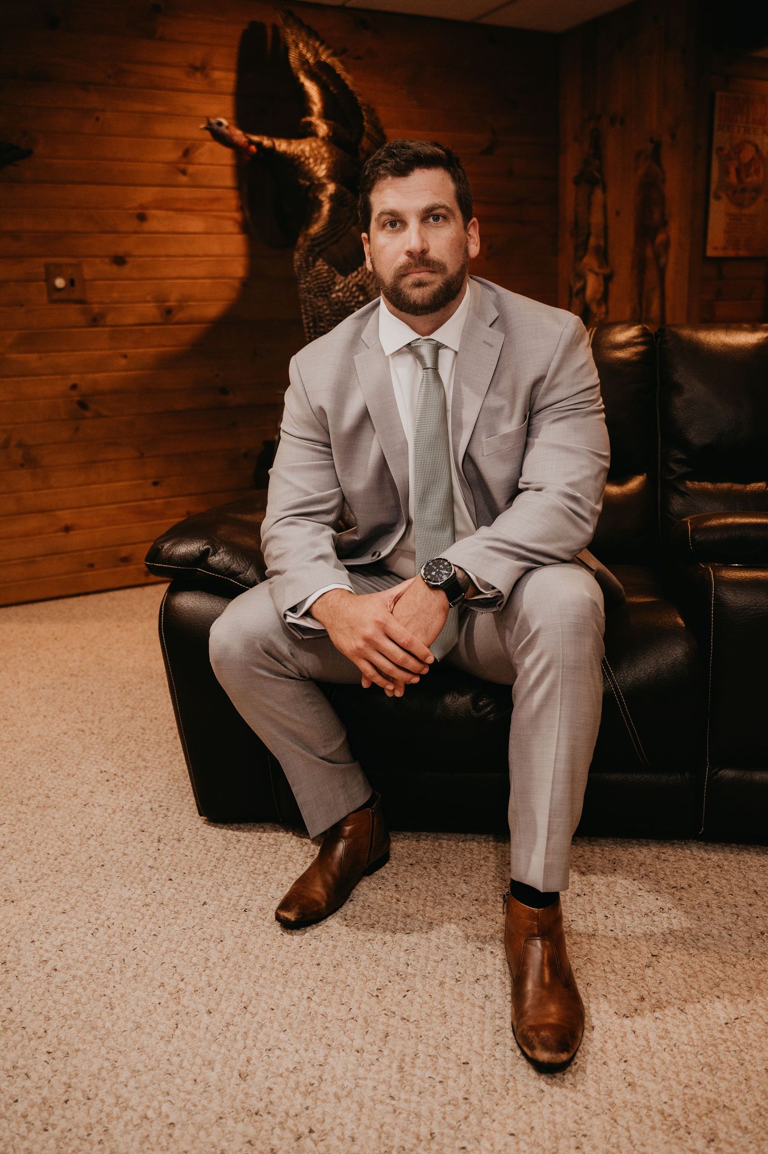 A person in a light gray suit and brown dress shoes sits on a black leather couch in a wood-paneled room.