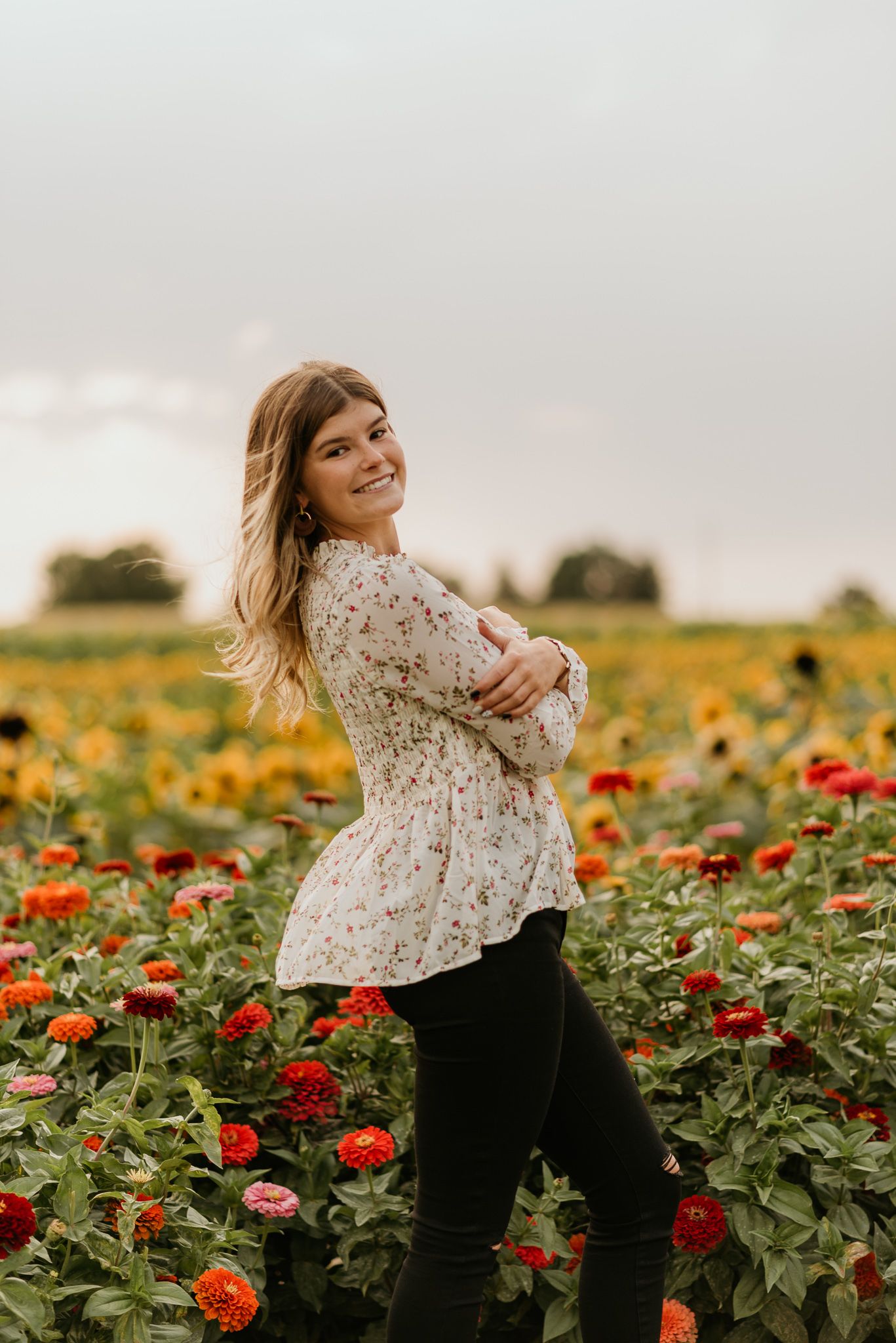 Woman in white lace top poses in a colorful garden filled with vibrant red and orange zinnias on an overcast day.