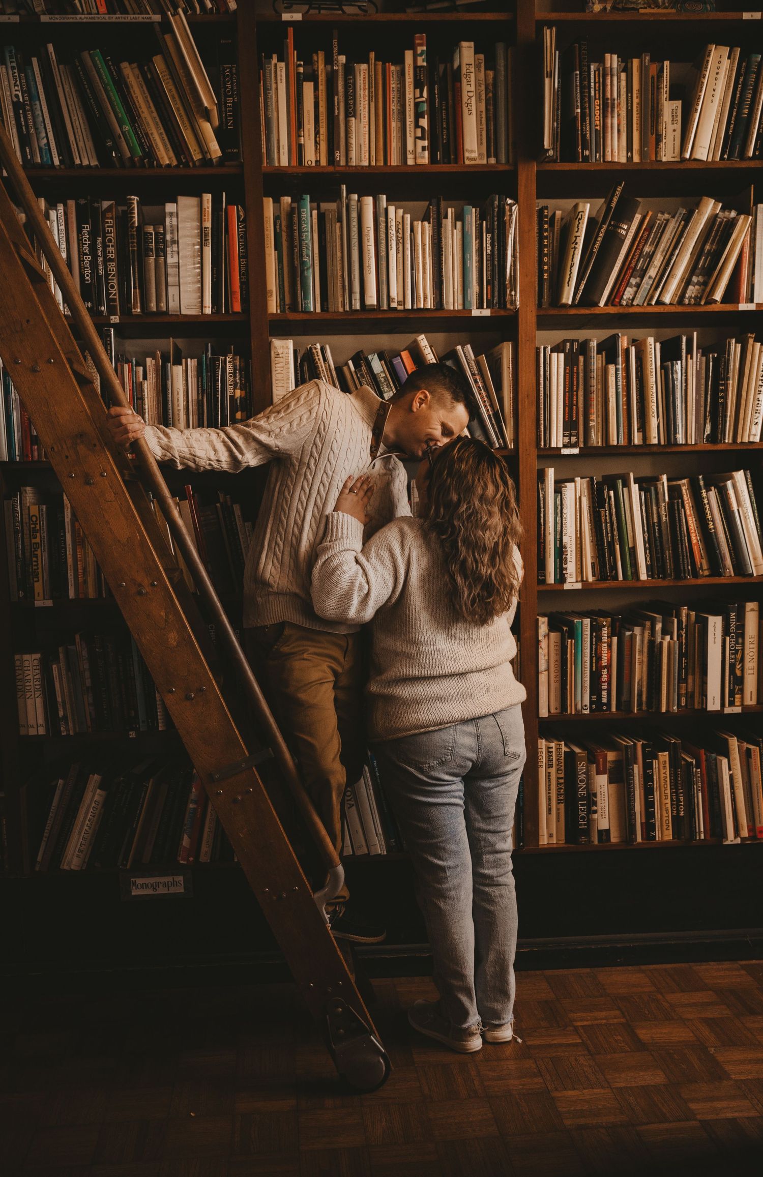 A person wearing white and grey clothing browses books on wooden library shelves in a dimly lit room.