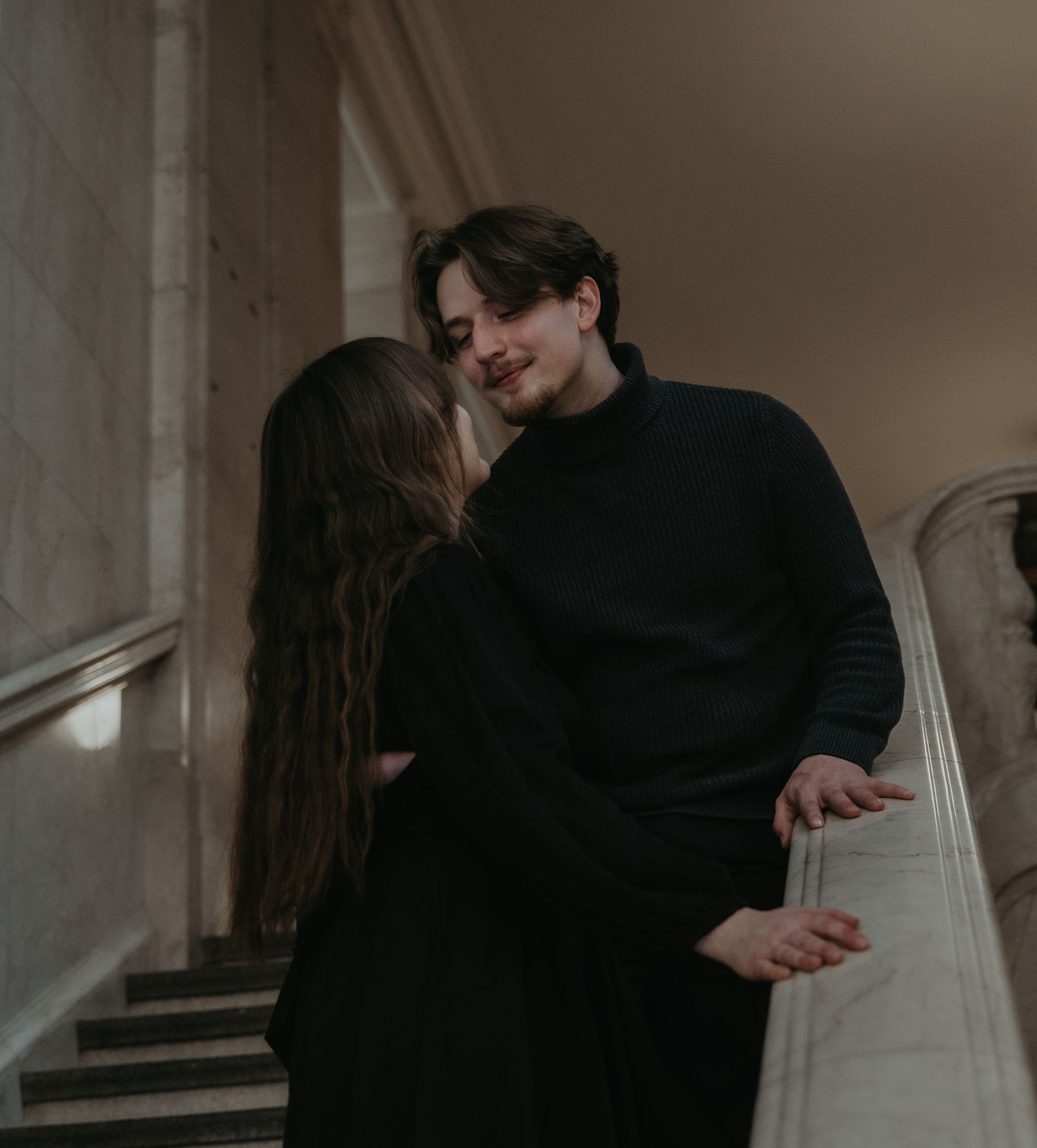 A couple embraces intimately in a dimly lit interior space with soft lighting and shadows.