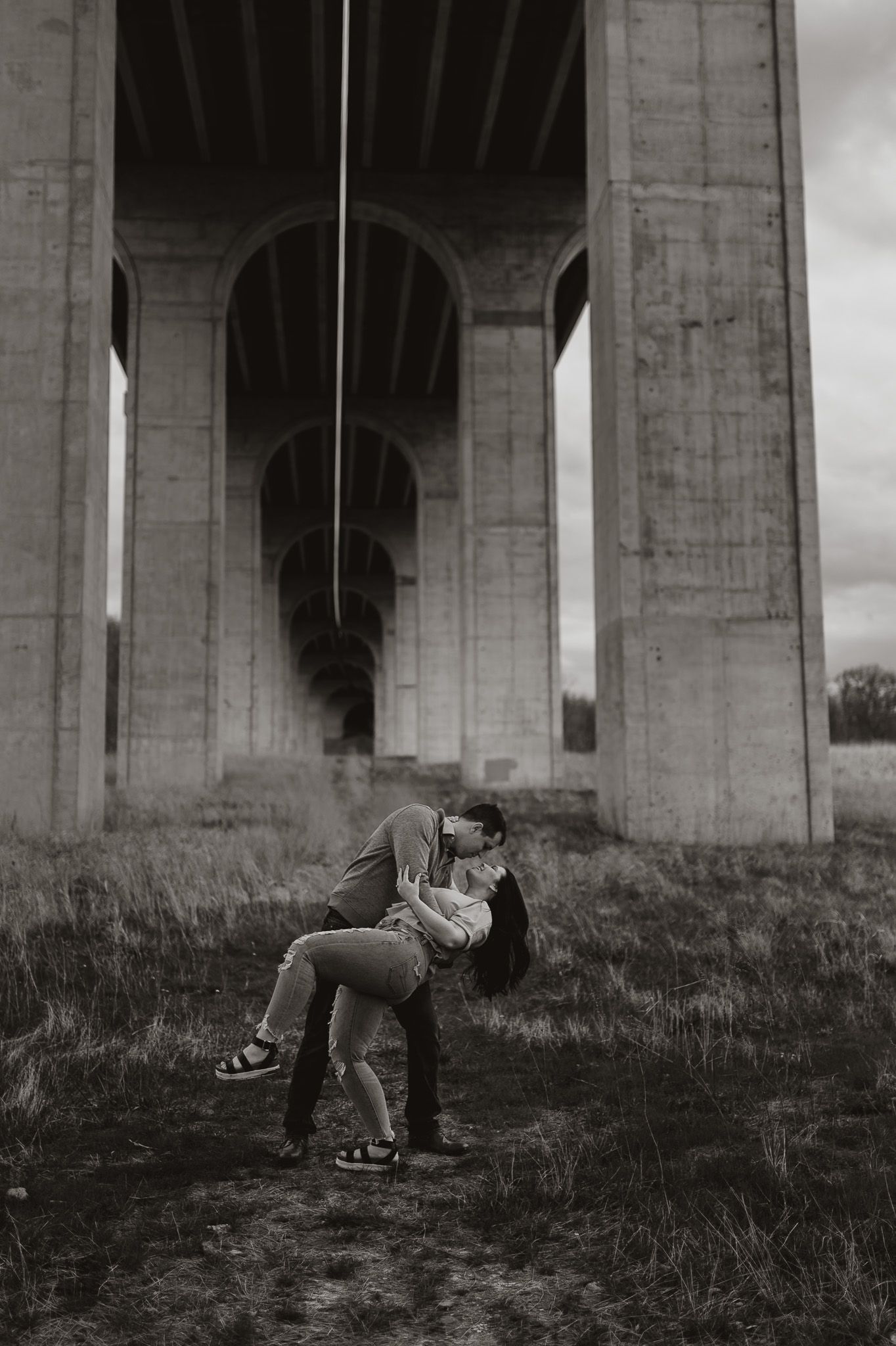Two people share an intimate dance-like pose beneath a concrete overpass.