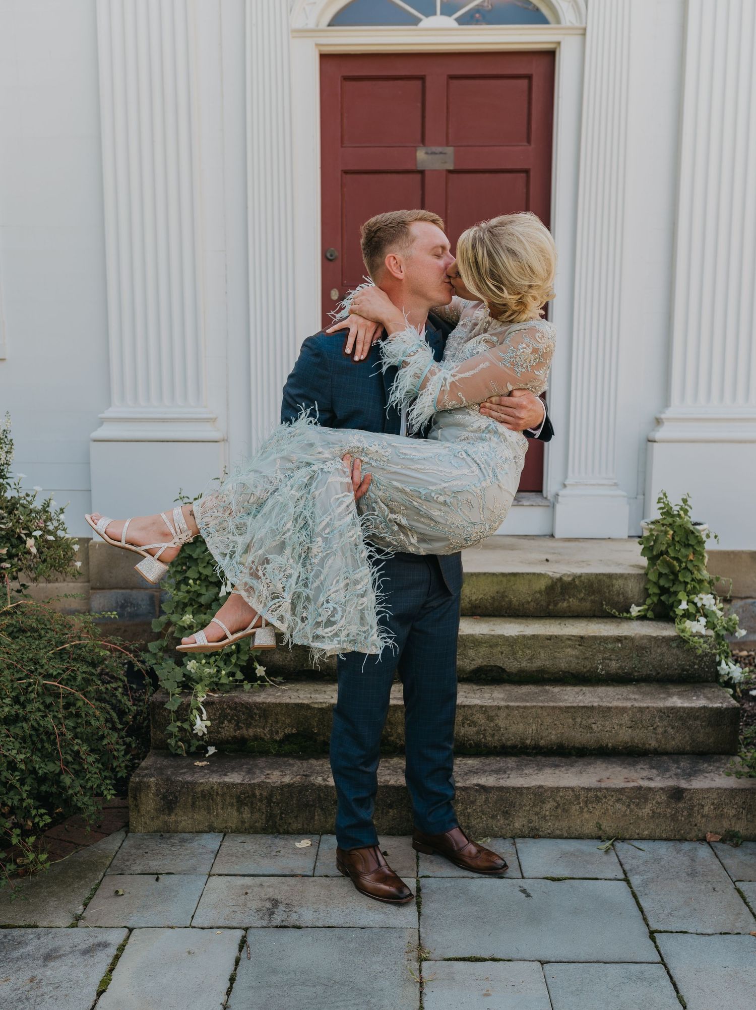 A couple shares a romantic moment in front of a red door as he carries her in her lace wedding dress.