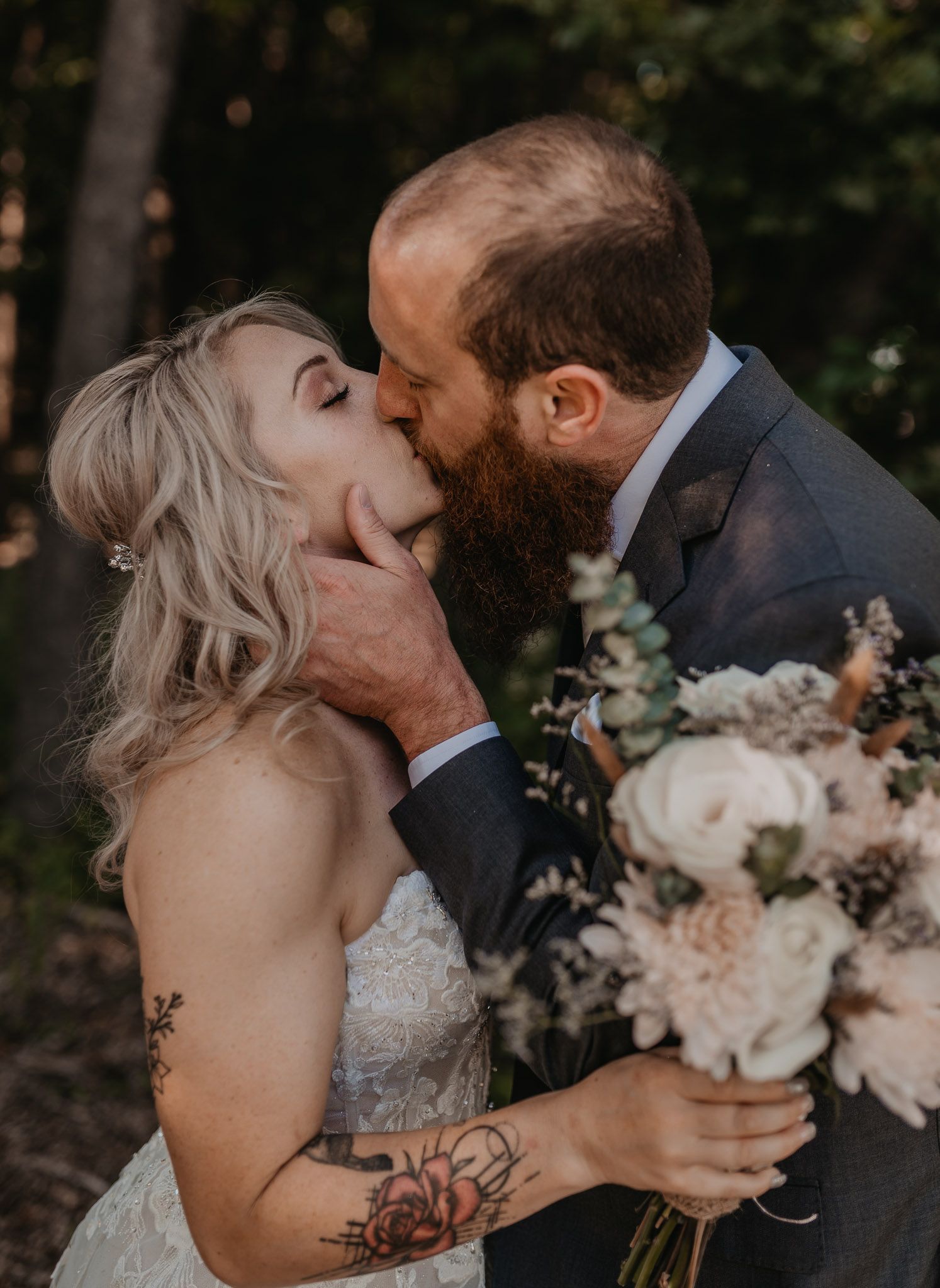 Passionate wedding kiss between bride and bearded groom while holding romantic floral bouquet.