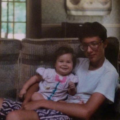 A man with glasses sits on a wicker couch holding a smiling toddler girl in a floral outfit in a cozy home setting.