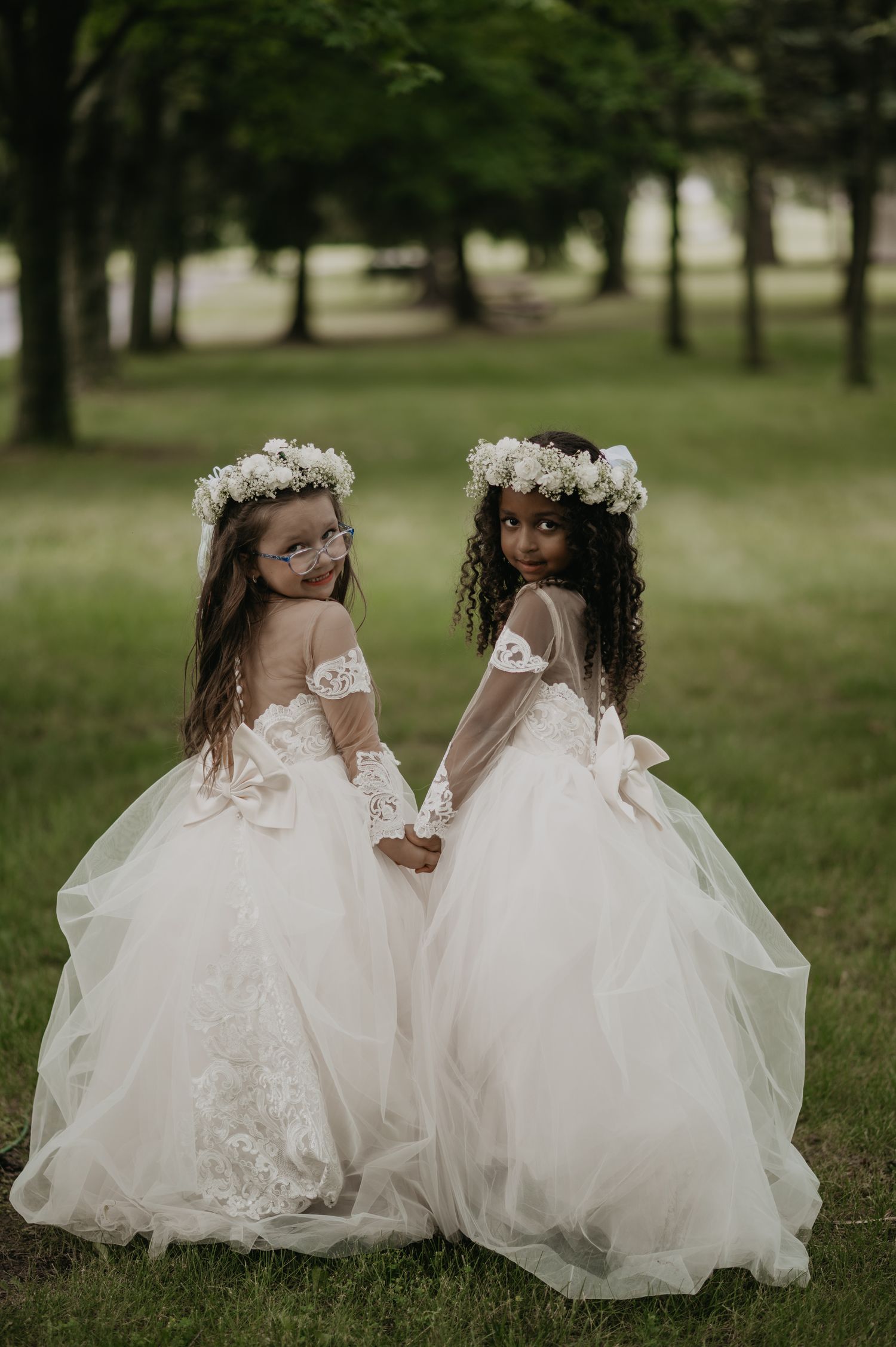 Two flower girls in matching white tulle dresses and floral crowns hold hands in a dreamy garden setting.