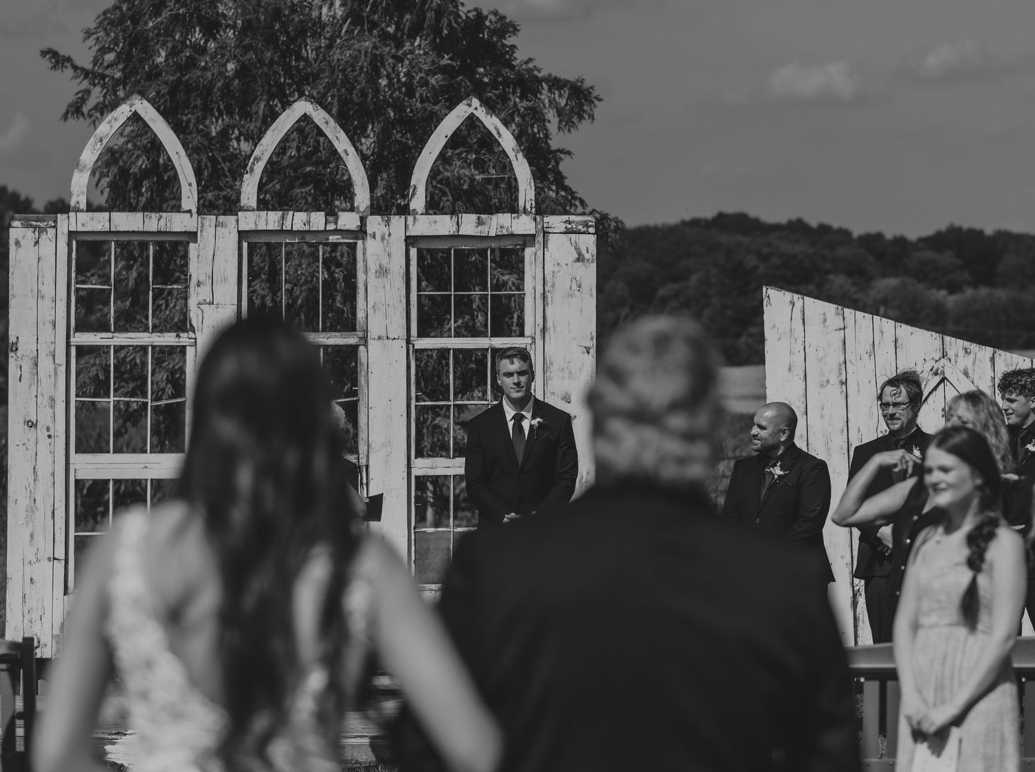 Outdoor wedding ceremony takes place in front of rustic gothic-style window frames against cloudy sky.