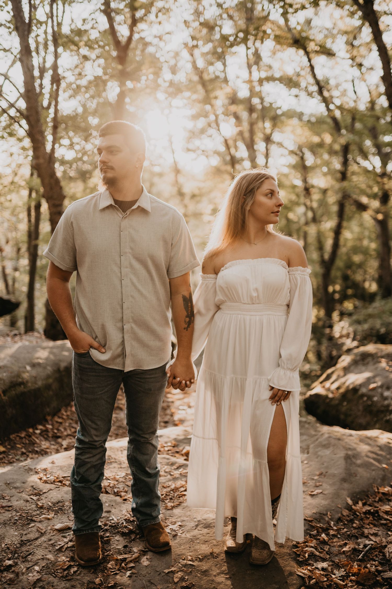 A couple walks hand in hand through a wooded area wearing white dress and casual attire.