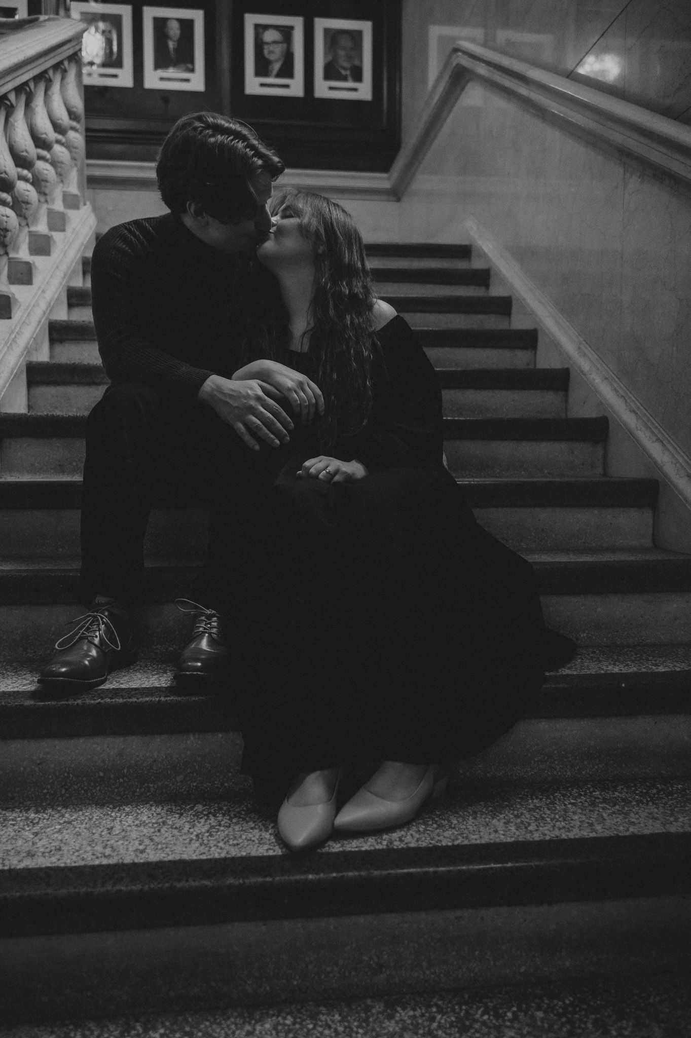 A romantic black and white photograph of a couple sitting intimately on a staircase with framed portraits on the wall.