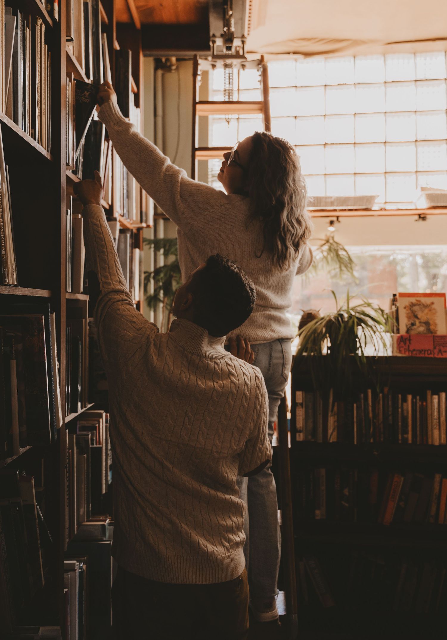 Silhouetted figure in a cozy room reaches up to grab a book from a tall wooden bookshelf at sunset.