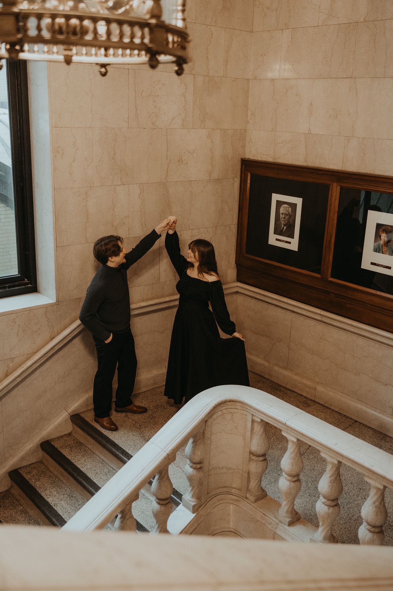 Two people in formal attire hang artwork on a beige wall along an elegant staircase in a historic building.