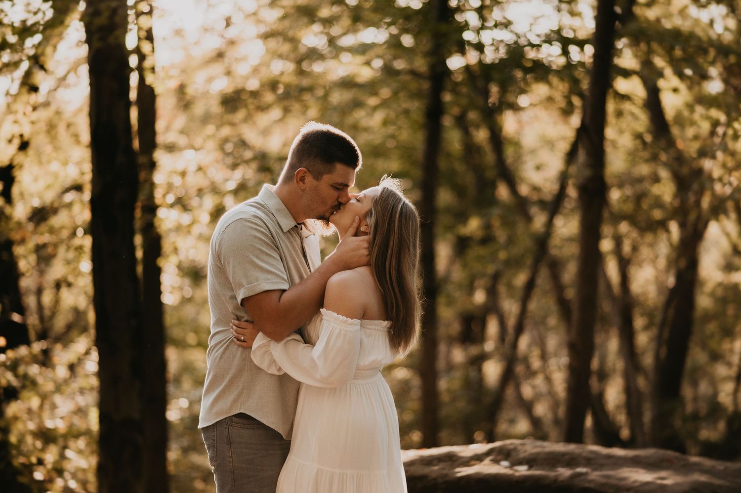 A romantic couple shares an intimate moment in a sunlit forest during a golden hour photo session.