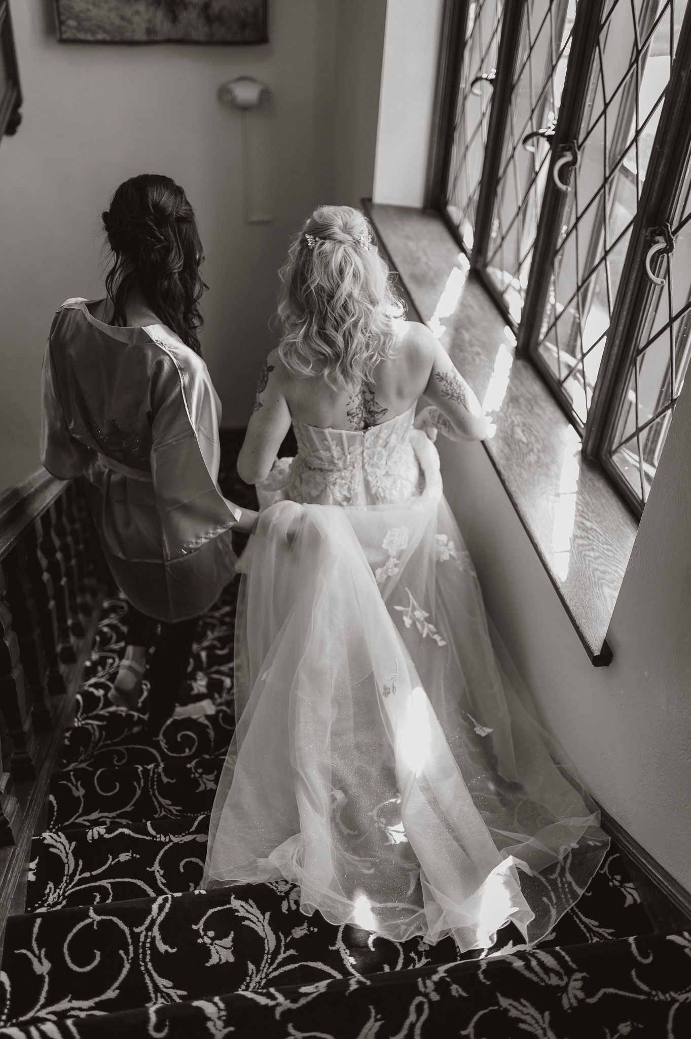 Black and white photo of bride and attendant looking out ornate windows in wedding attire.