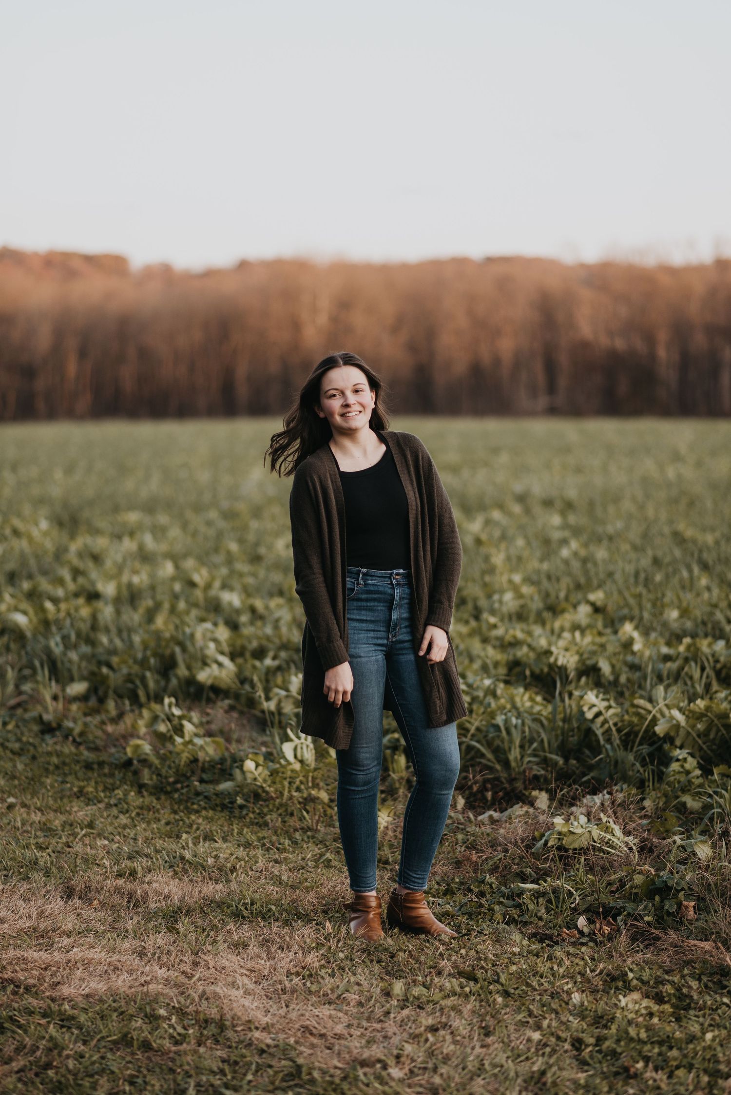 A person stands on the edge of a green field at sunset wearing a long cardigan and jeans with autumn trees in background.