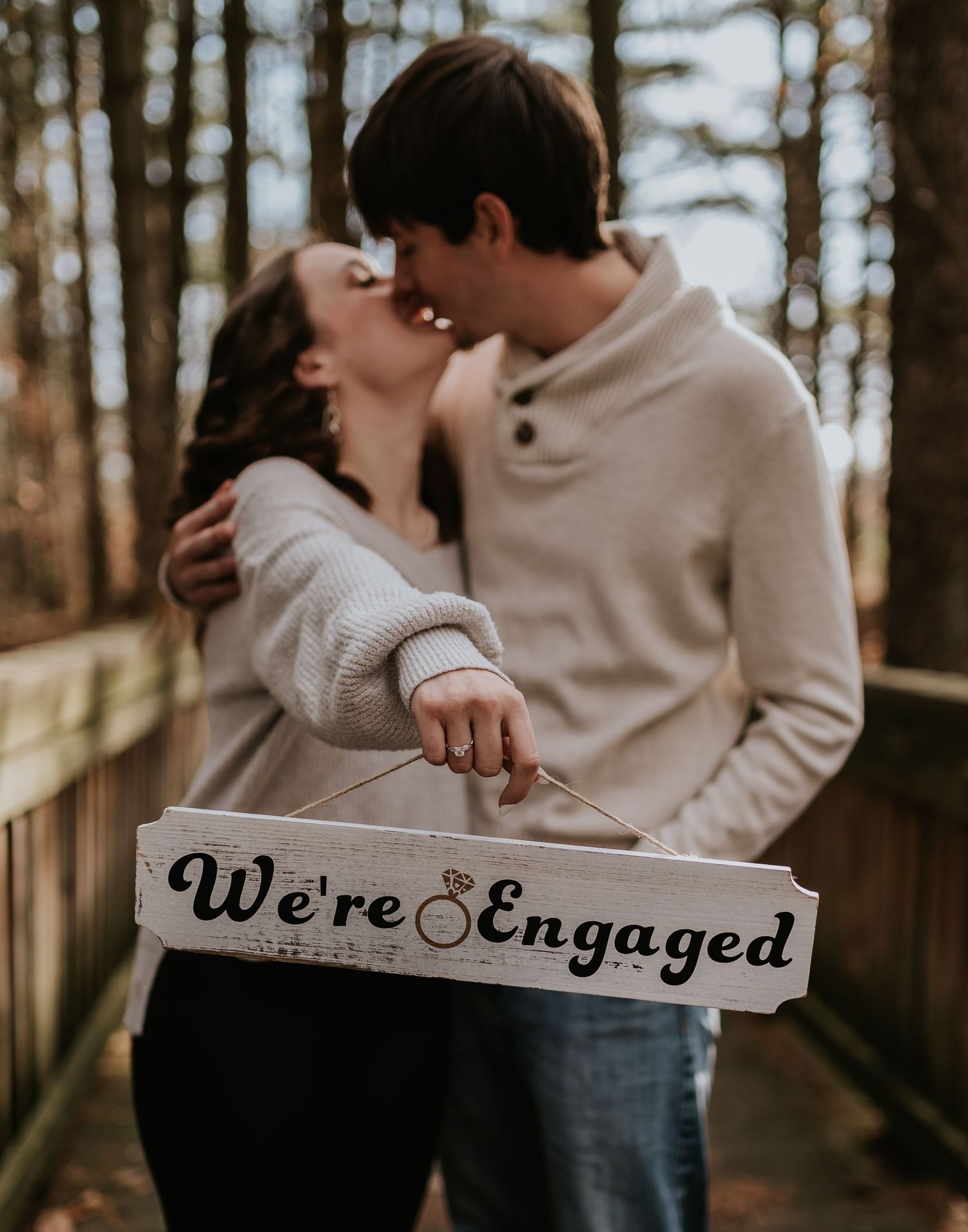 Engaged couple sharing intimate moment while displaying wooden announcement sign.