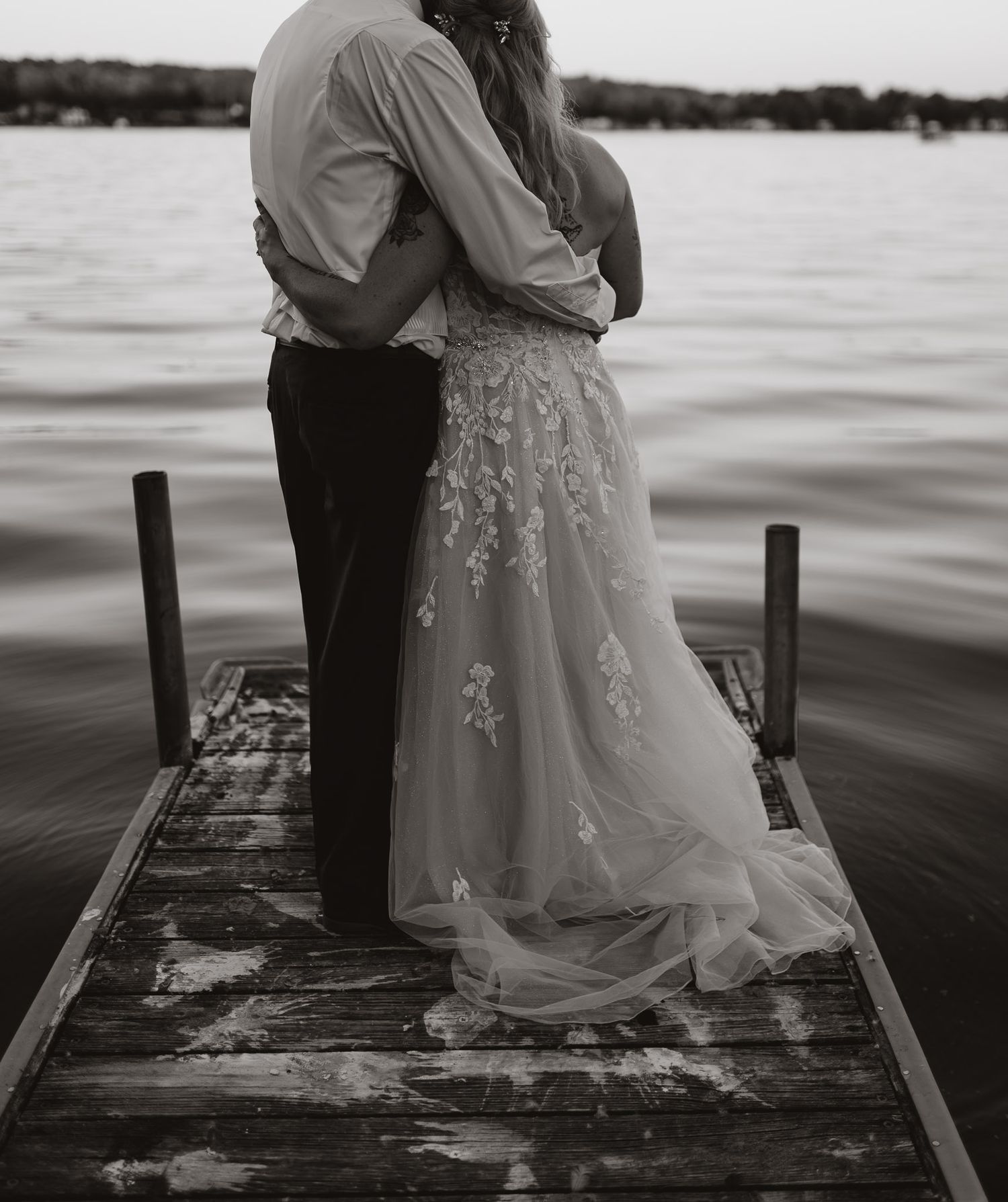 Black and white photo of newlyweds embracing on a dock with water in the background.