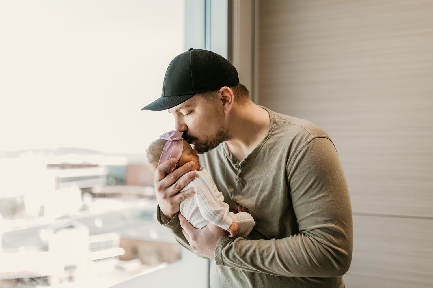Father tenderly kissing newborn baby by window in soft natural light.