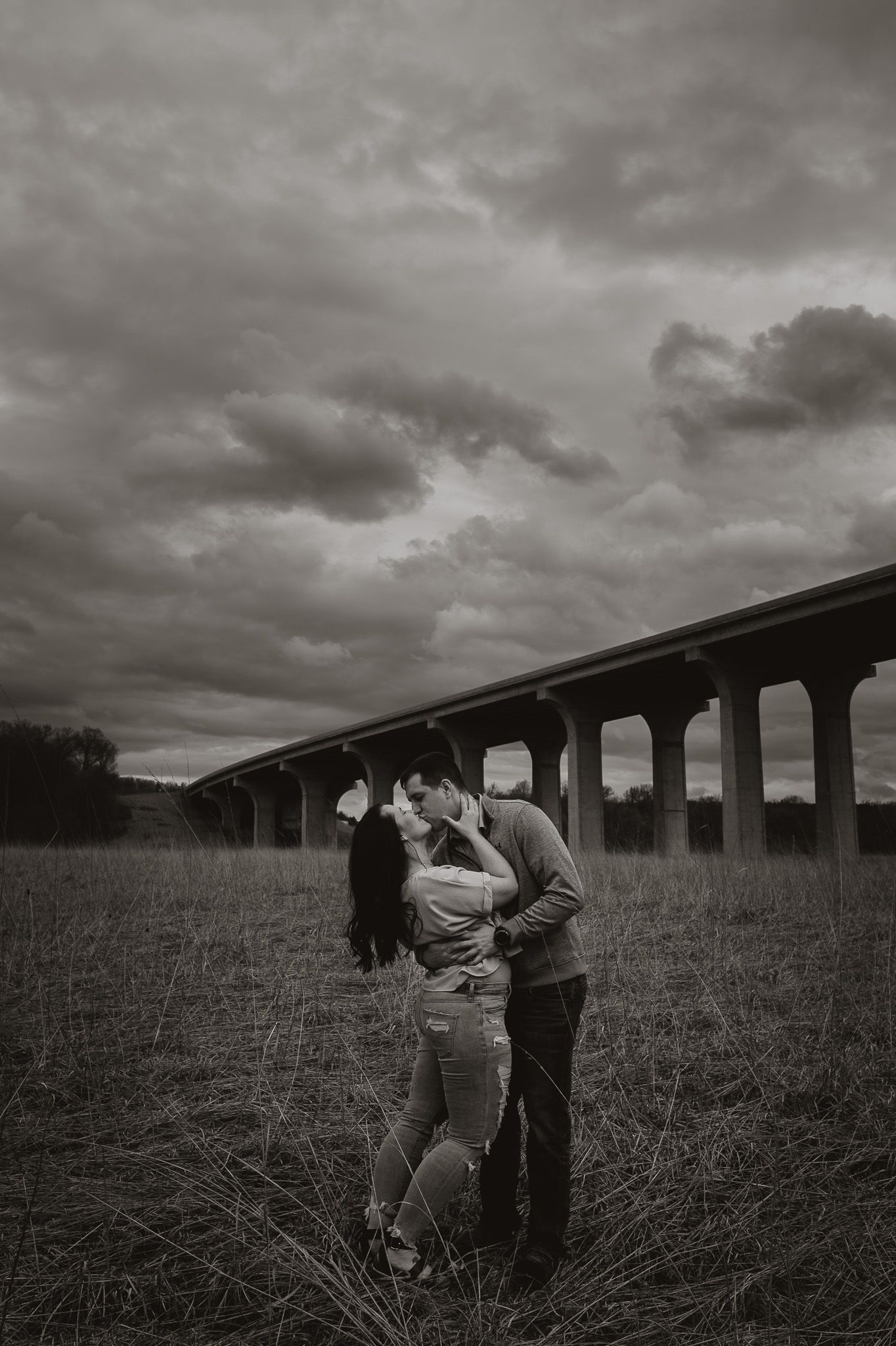 A romantic black and white portrait of a couple embracing in a field beside a bridge.