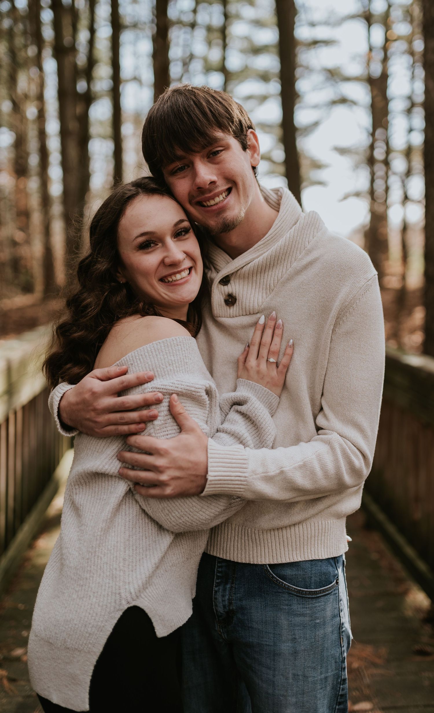 Couple in cozy cream sweaters embracing on a wooden bridge in an autumn forest setting.