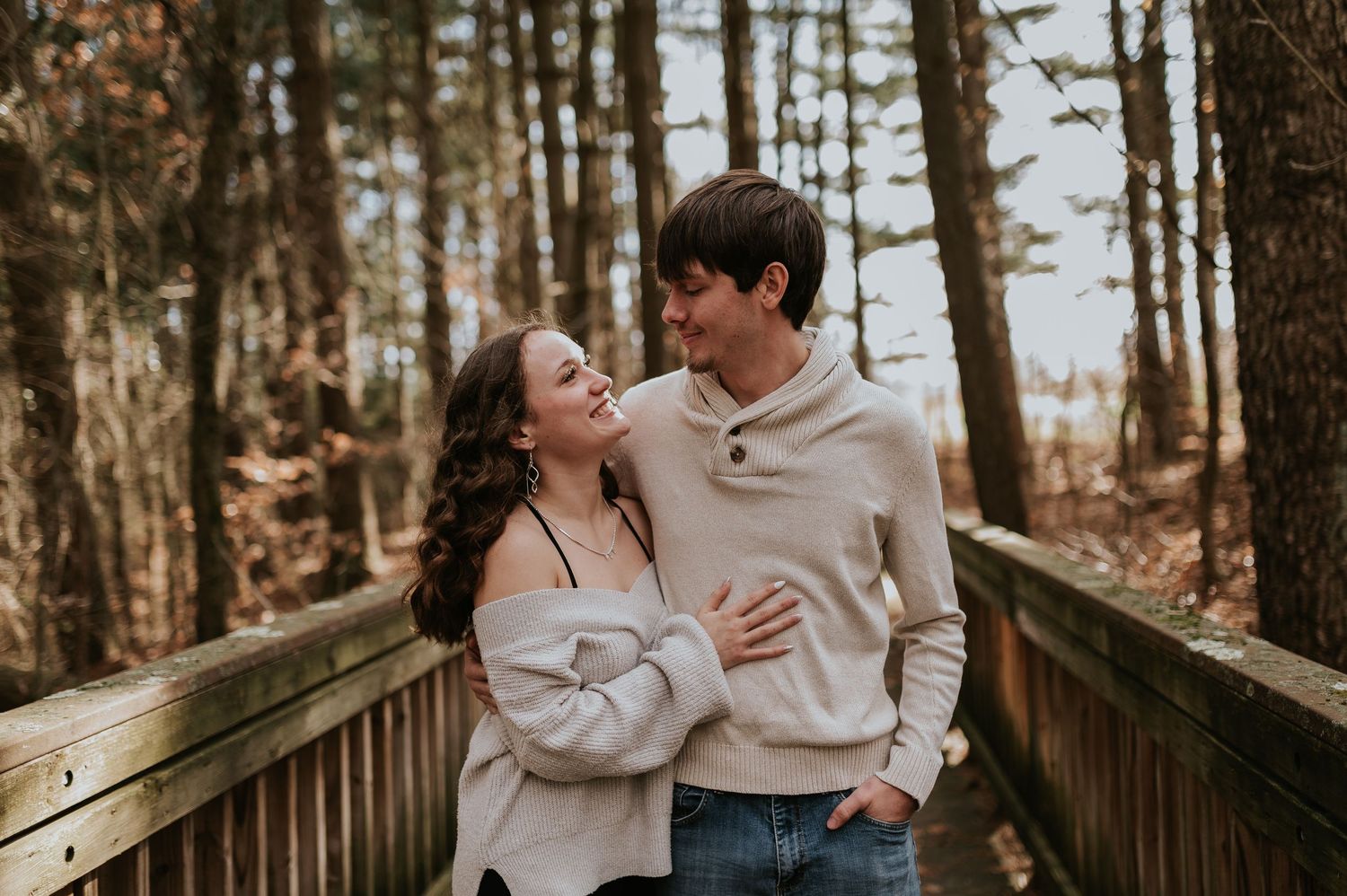 A couple shares a joyful moment together on a wooden boardwalk surrounded by tall trees in autumn.
