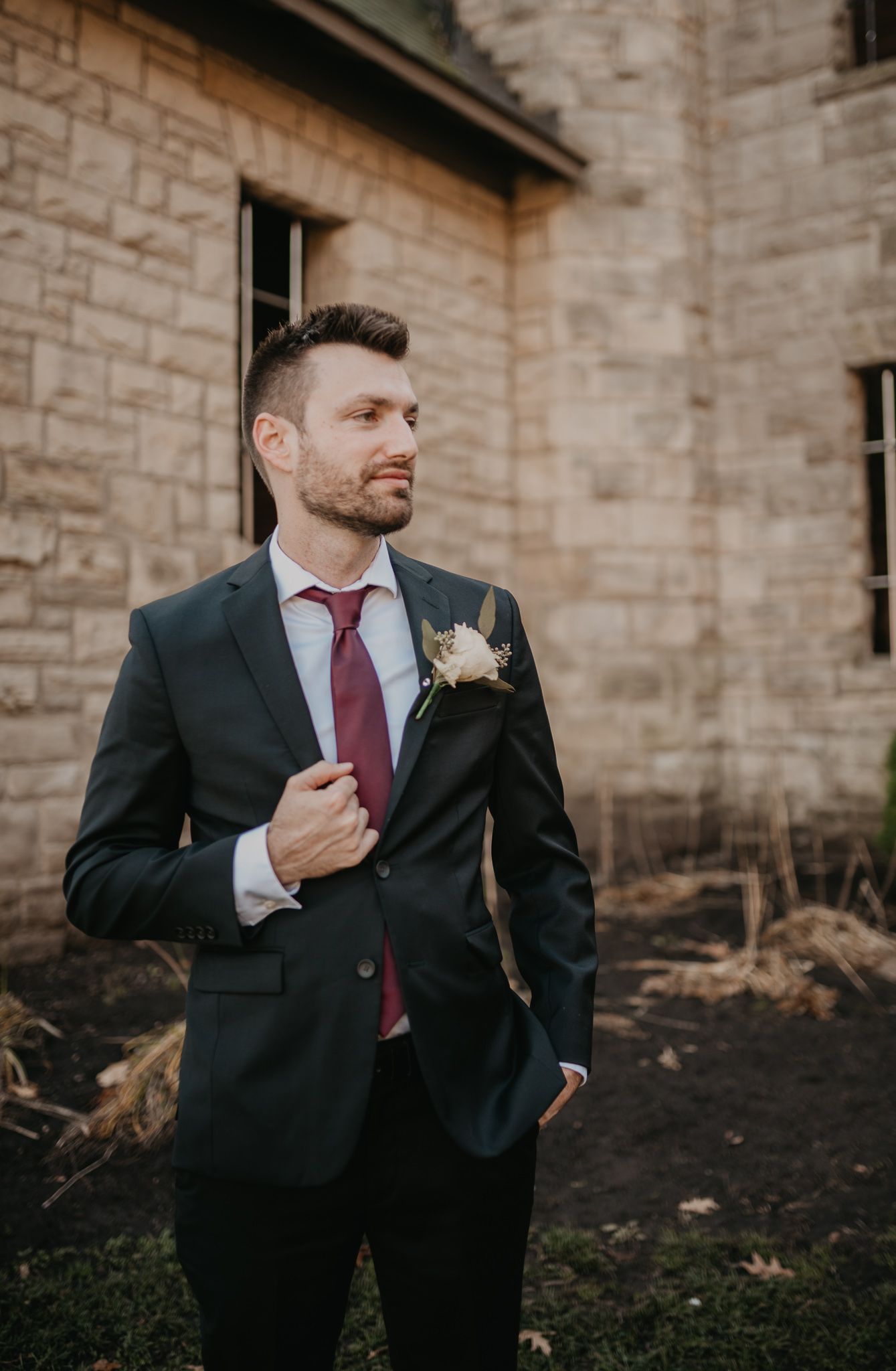 Groom in dark suit with burgundy tie poses confidently against stone building exterior.