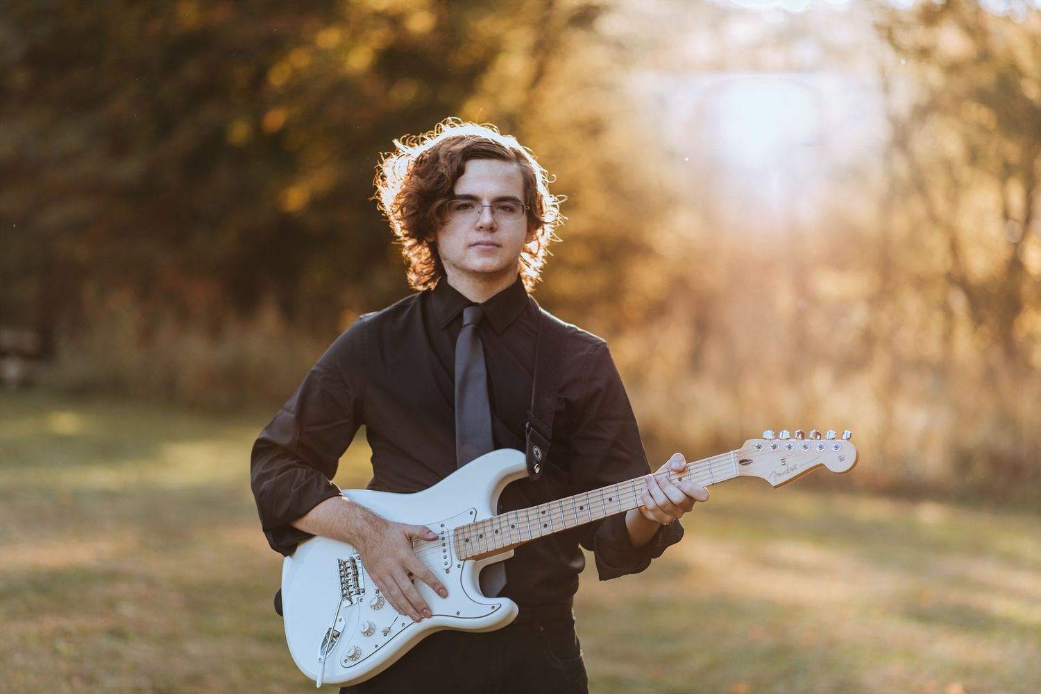 A person in a black coat plays a white Fender Stratocaster guitar in a golden sunlit outdoor setting during autumn.