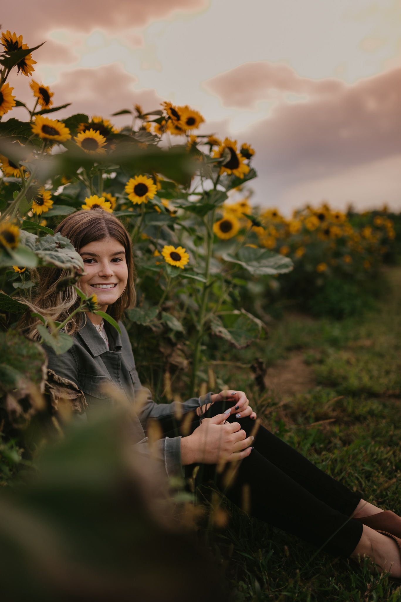A soft sunset glow illuminates a vibrant sunflower field where someone sits with a camera among the tall yellow blooms.