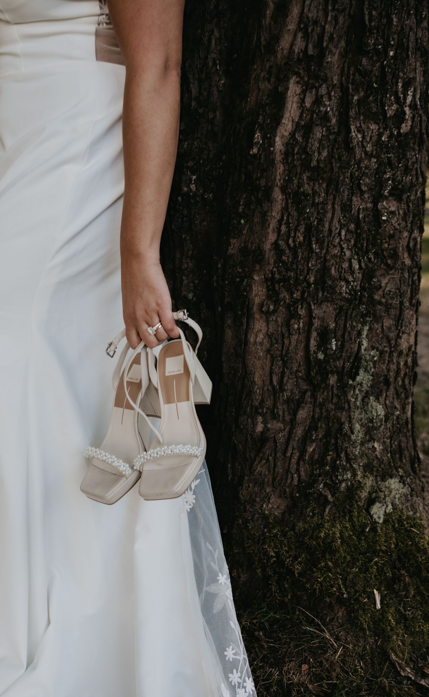 A pair of elegant beige wedding heels held against a white dress and dark tree trunk background.