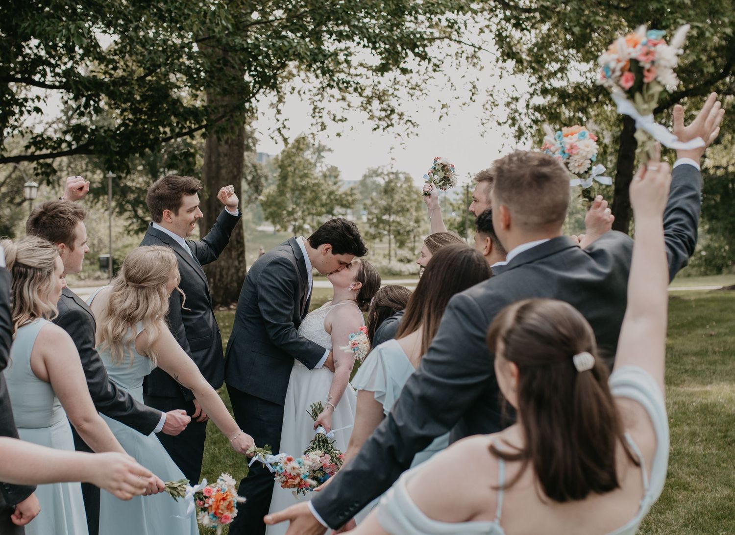 A joyous celebration at an outdoor wedding ceremony with guests cheering and raising floral bouquets.