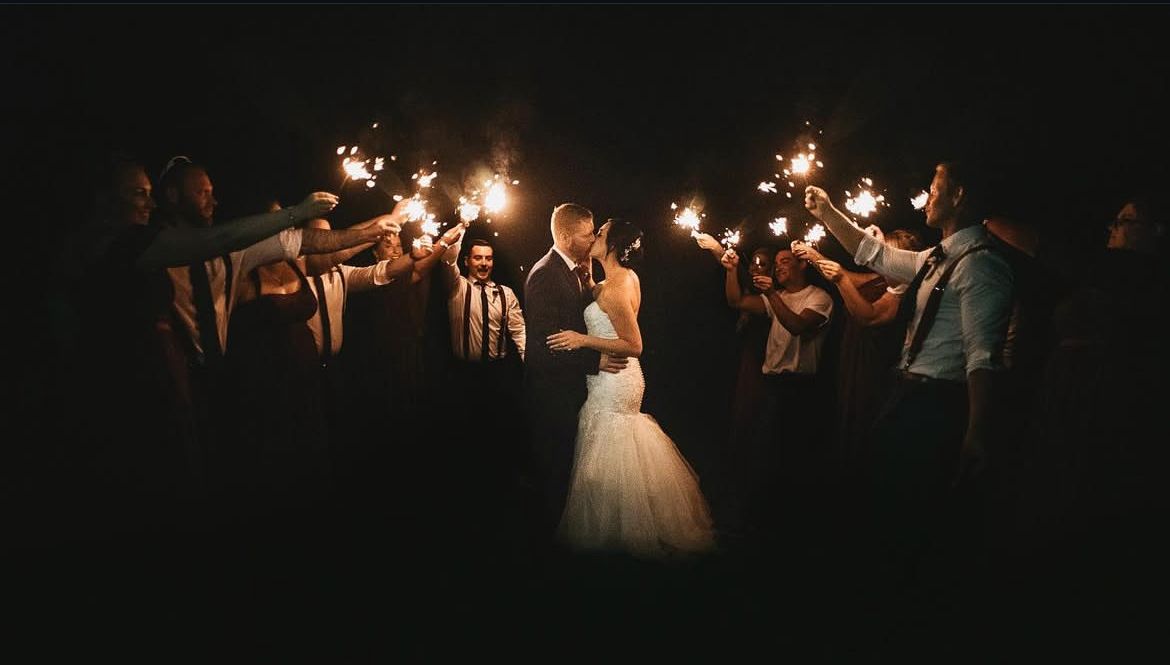 Newlyweds share an intimate moment surrounded by sparkler-holding guests at night.