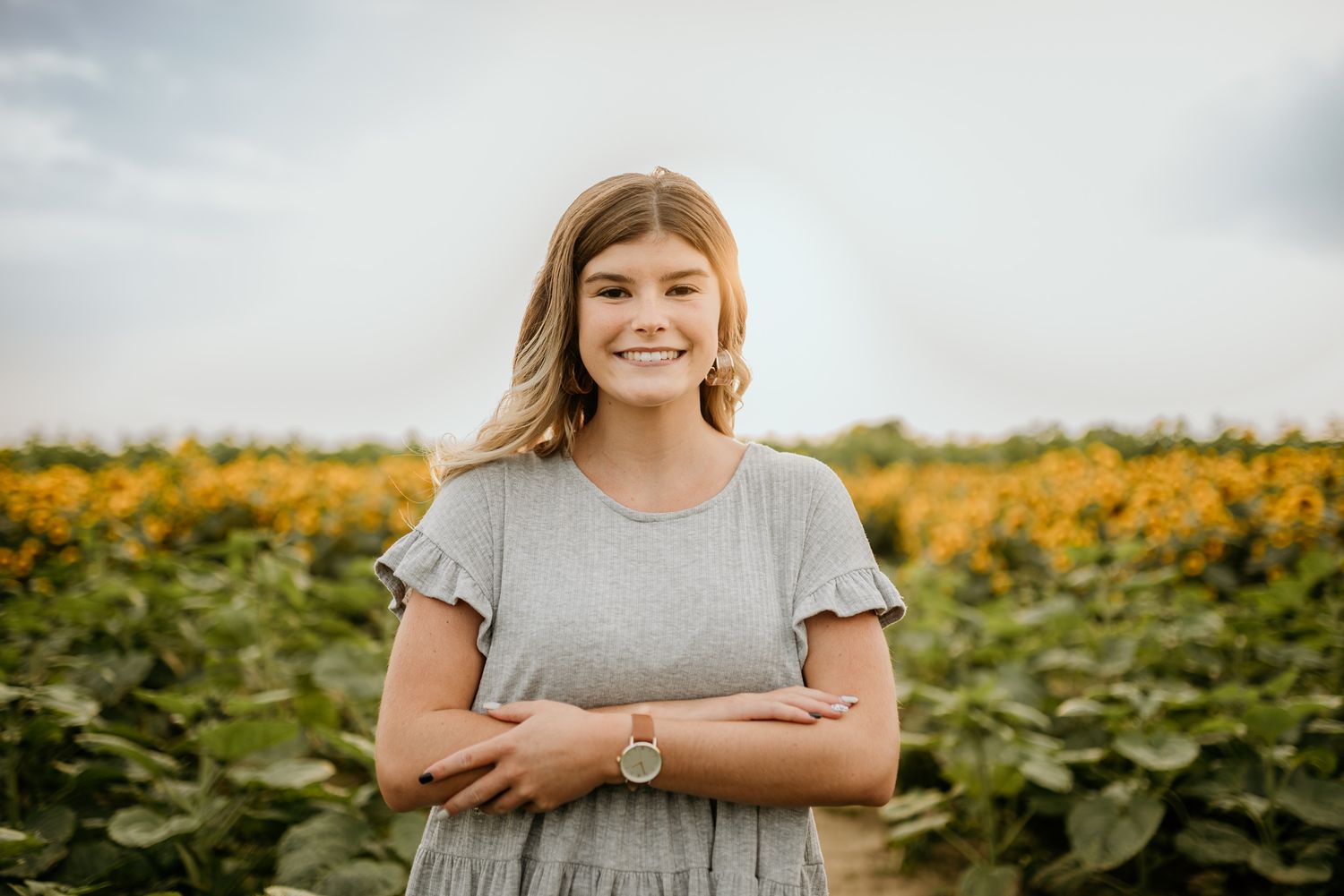 Person in gray dress smiling in front of vibrant sunflower field on cloudy summer day.