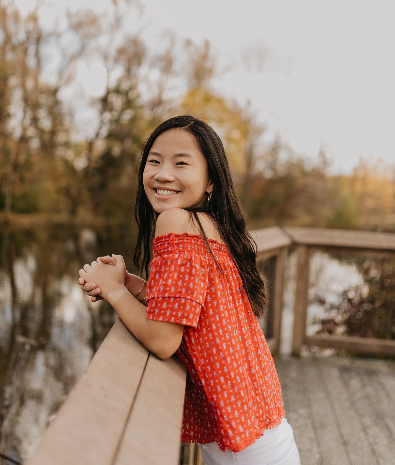 Candid moment on wooden deck wearing coral off-shoulder top by peaceful pond.