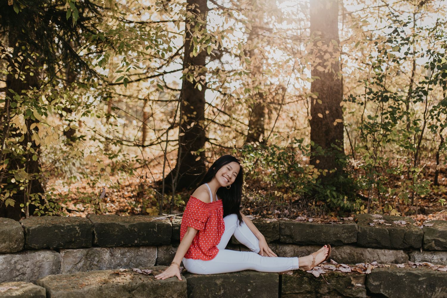 Relaxed pose in red top against sunlit forest background.