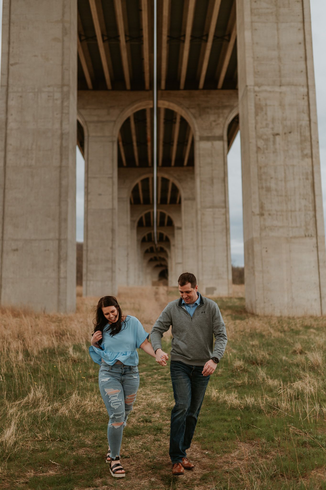 Two people playfully run down a grassy slope under a large concrete bridge structure.