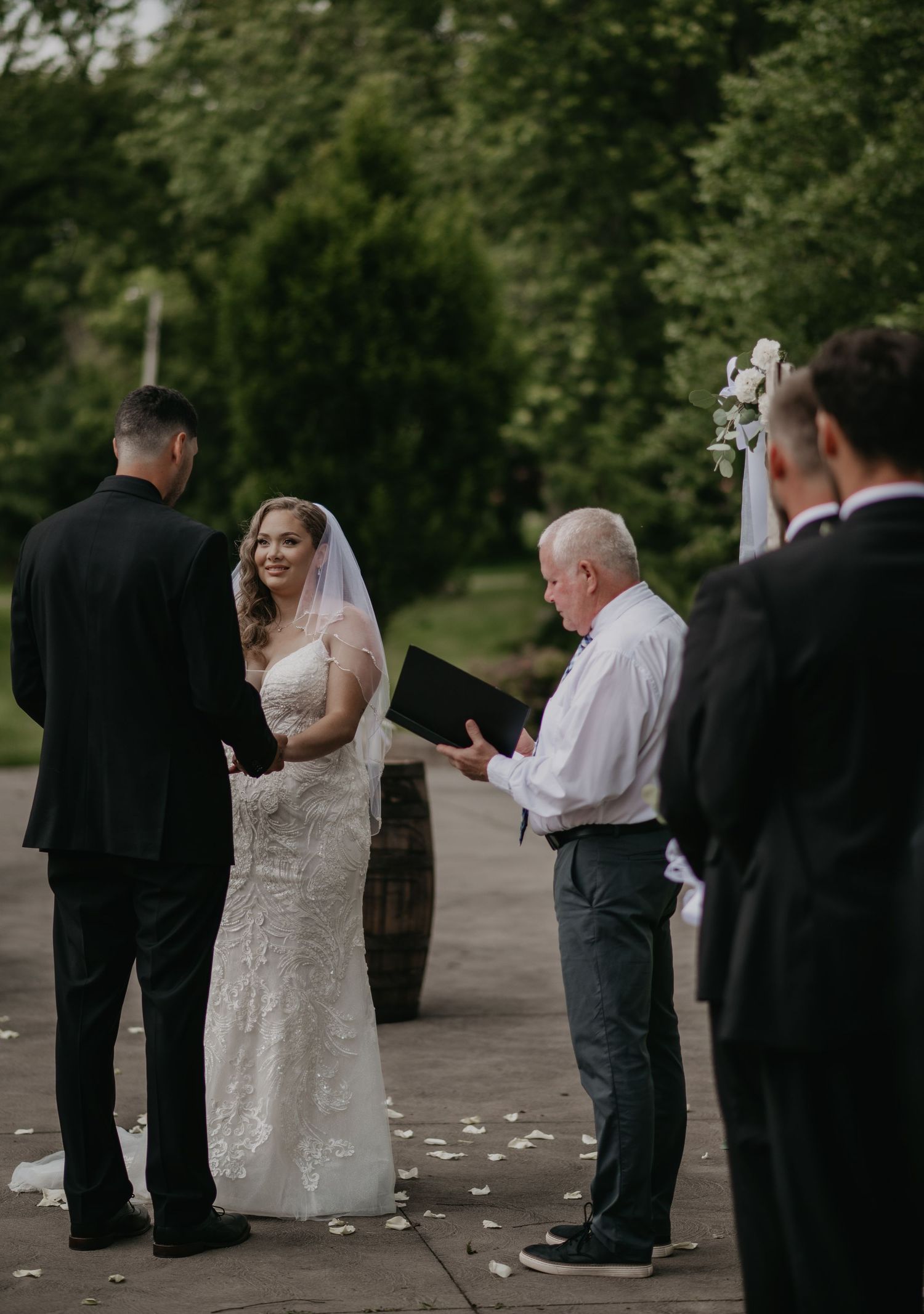 Wedding ceremony participants stand in a line outdoors surrounded by trees and scattered flower petals.