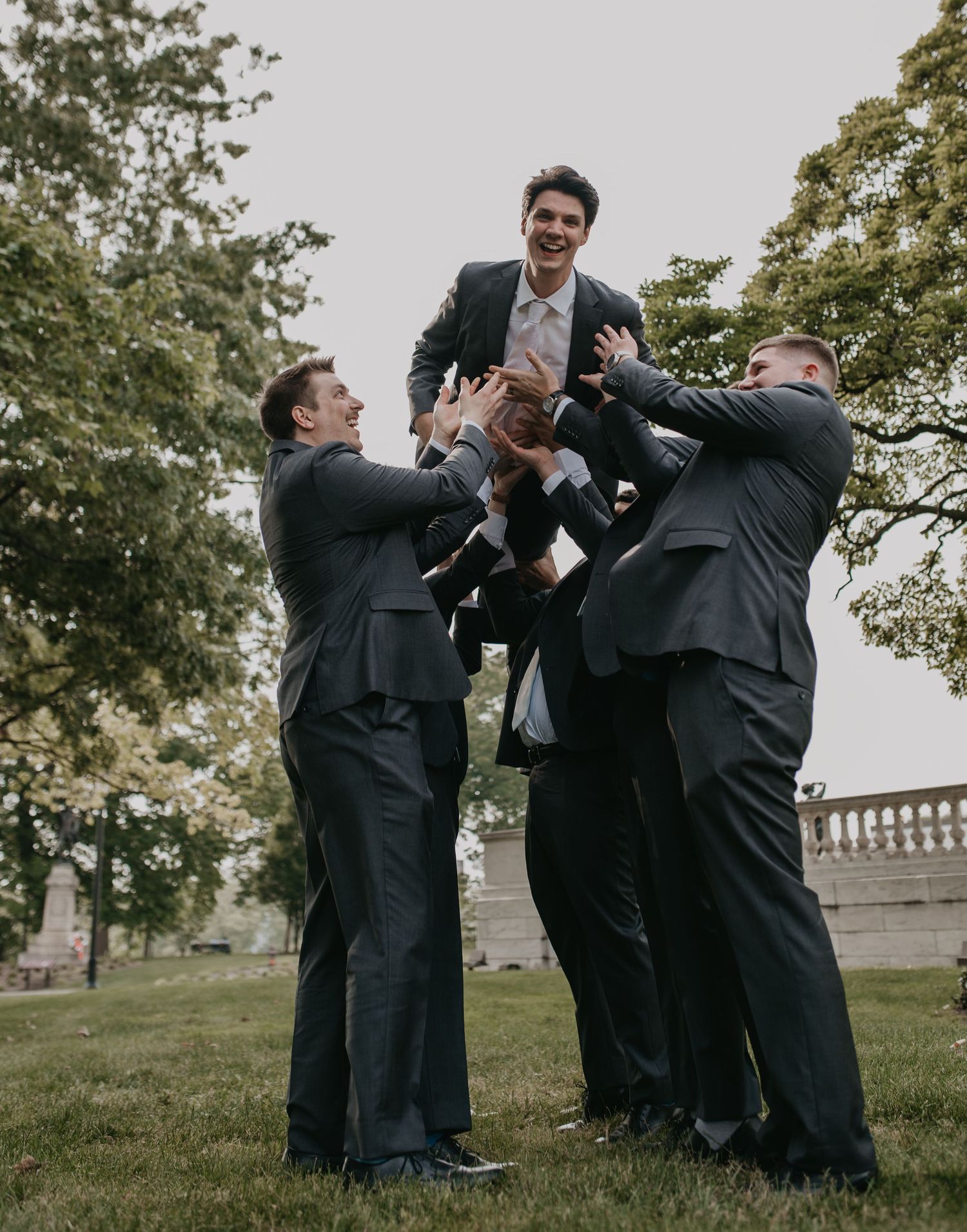 Groomsmen in black suits playfully spraying cologne outdoors during a wedding photoshoot.