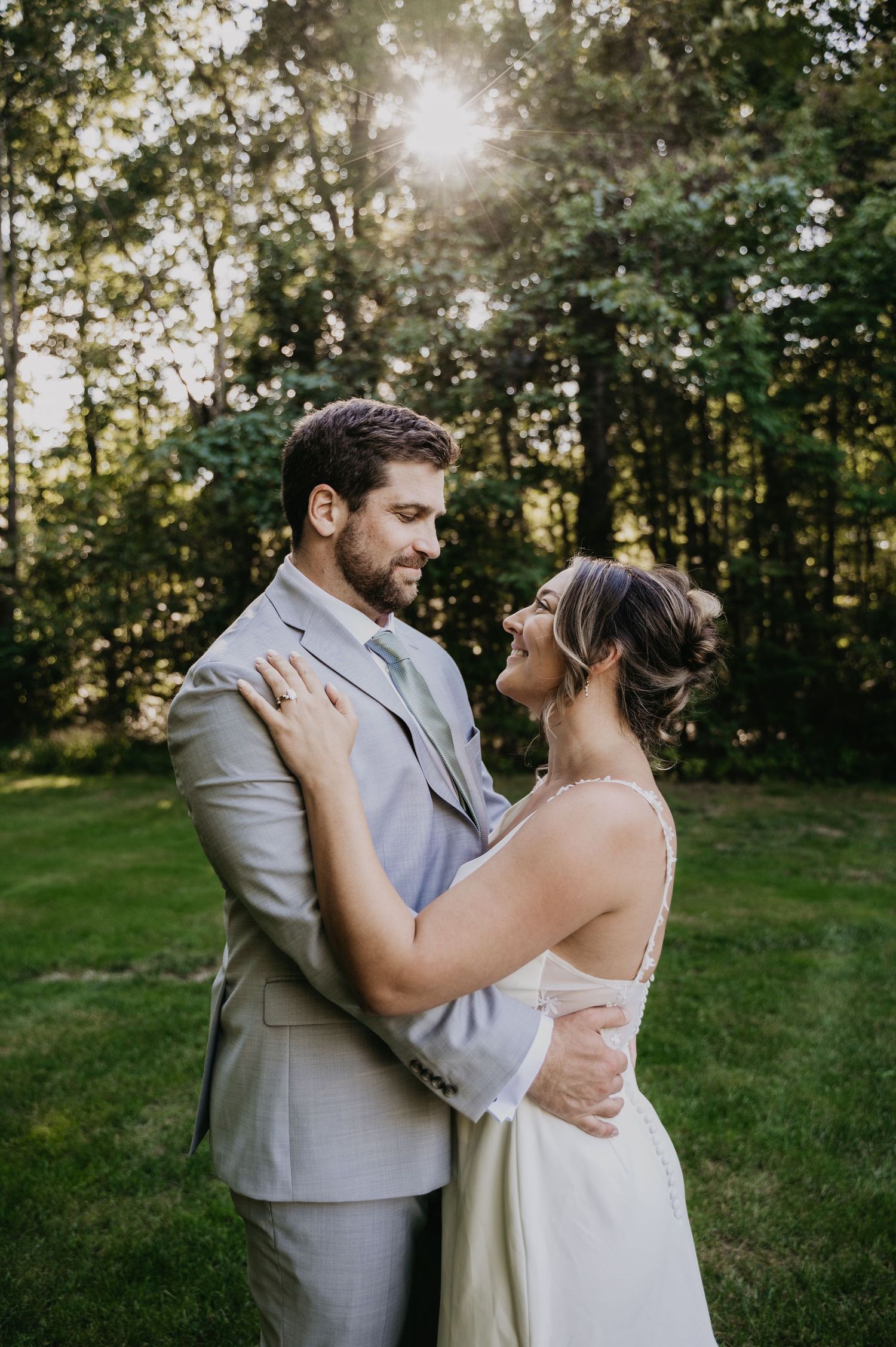 A romantic outdoor wedding portrait of a couple embracing in a sunlit wooded setting wearing gray suit and white dress.