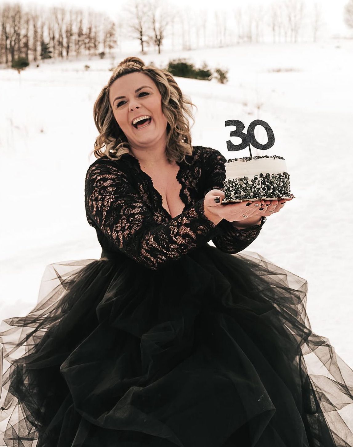 Woman wearing dramatic black tulle gown laughs joyfully in snowy winter landscape.