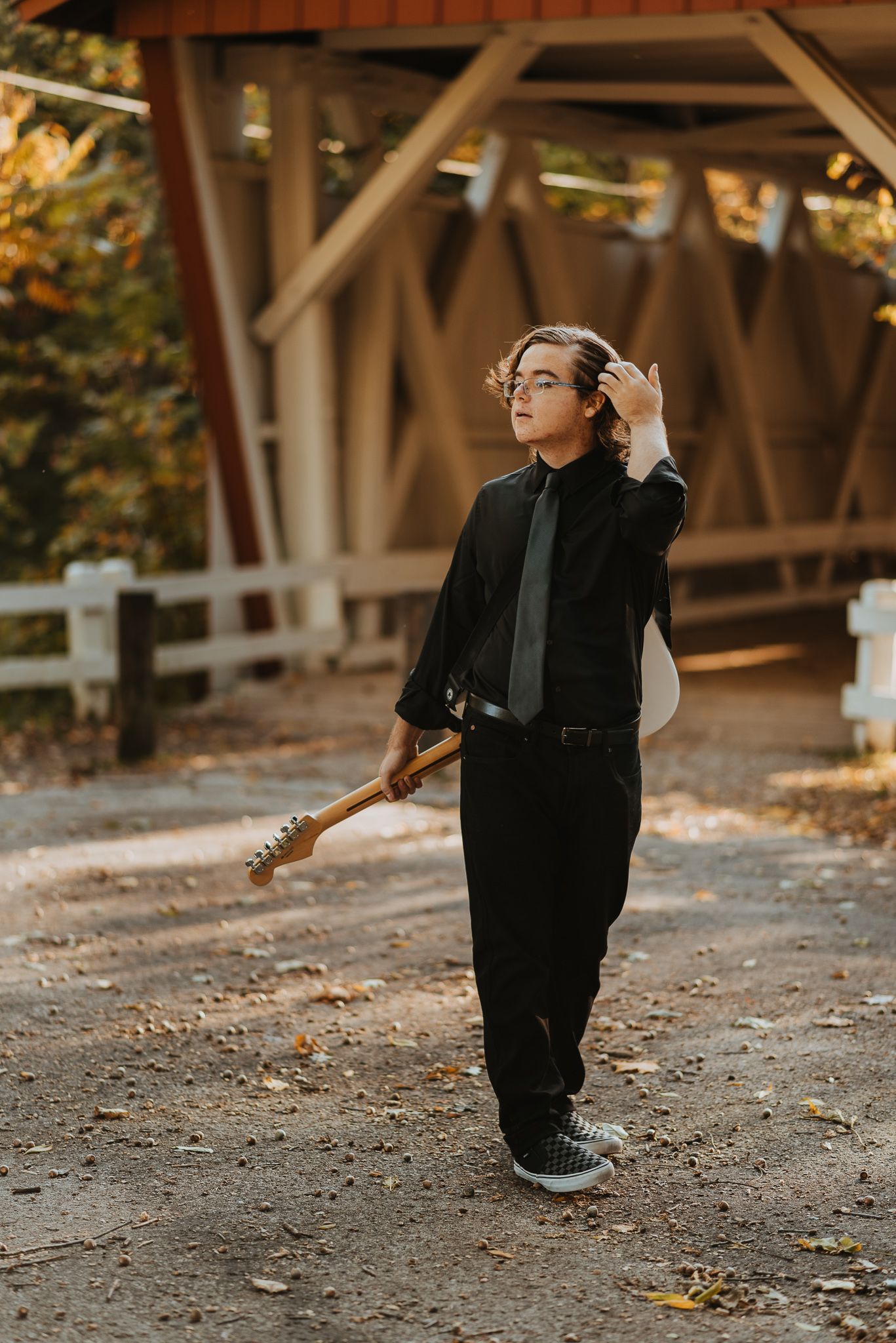 A musician in all black holds an electric guitar while standing on a dirt path beneath a wooden bridge structure.